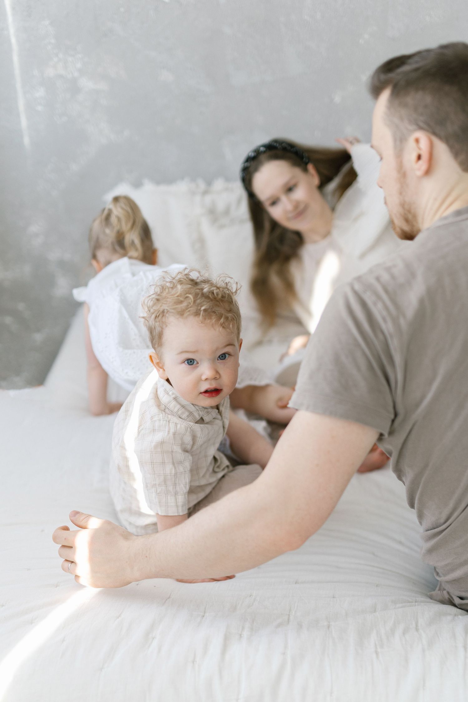 A happy family with two young children sitting together on a white bed in a bright, airy bedroom.