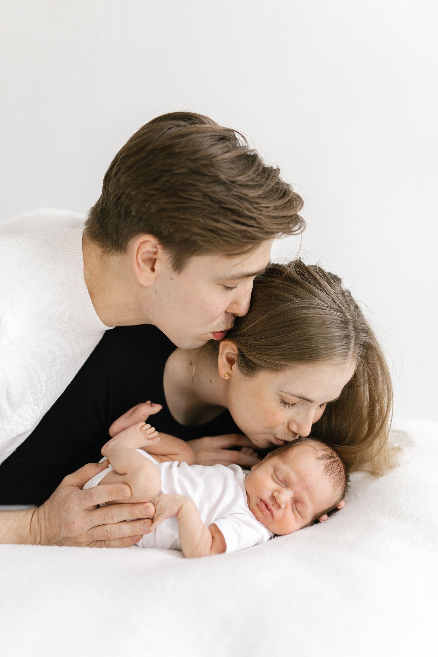 A couple tenderly kisses their sleeping newborn baby dressed in white, lying on a soft white blanket.