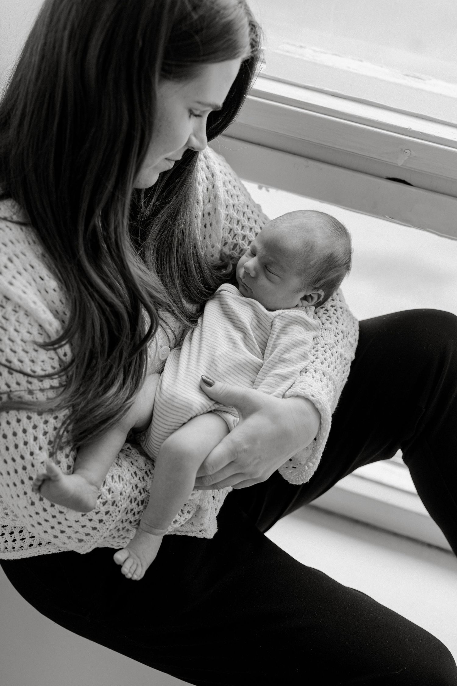 Mother in knit sweater holding sleeping newborn by window.