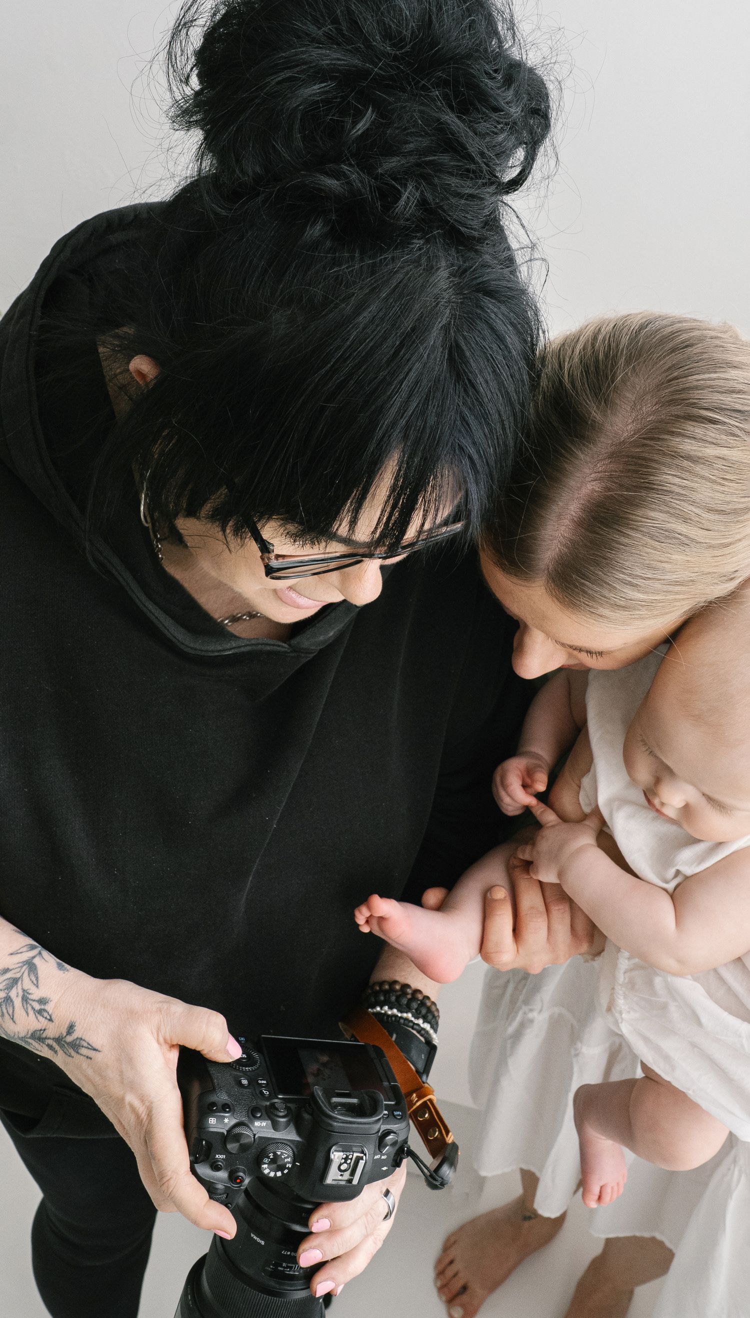 Photographer with black hair and tattoos holds a camera while a mother presents her newborn baby for a photo session.