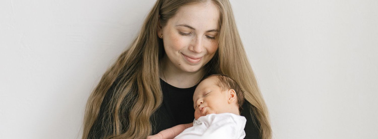 A young mother with long blonde hair tenderly holds her sleeping newborn baby dressed in white against a light background.