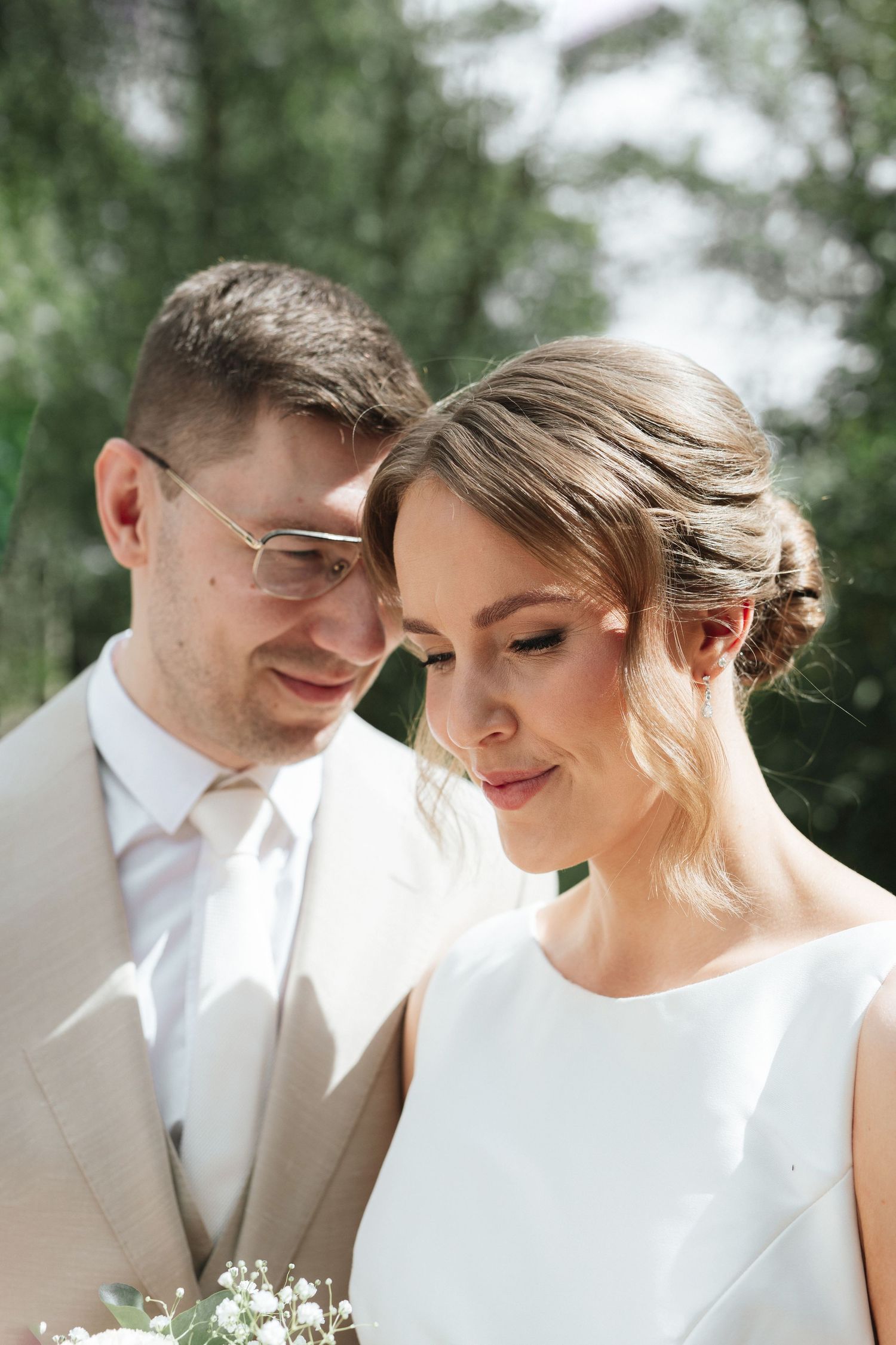 A bride and groom share a tender moment outdoors, with the groom in a beige suit and bride holding a white bouquet.