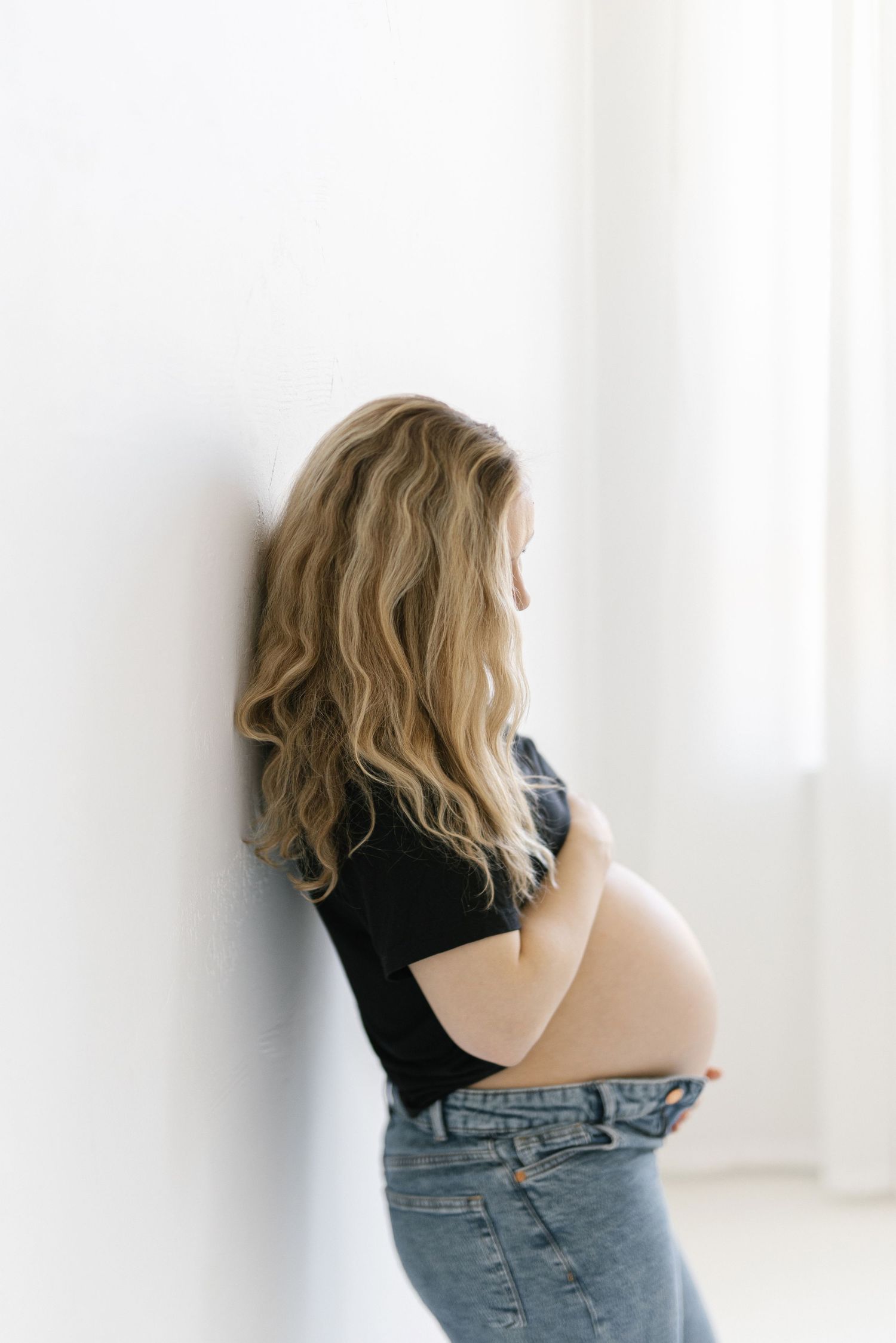 Pregnant woman with blonde wavy hair leaning against white wall, wearing black top and jeans, cradling baby bump.