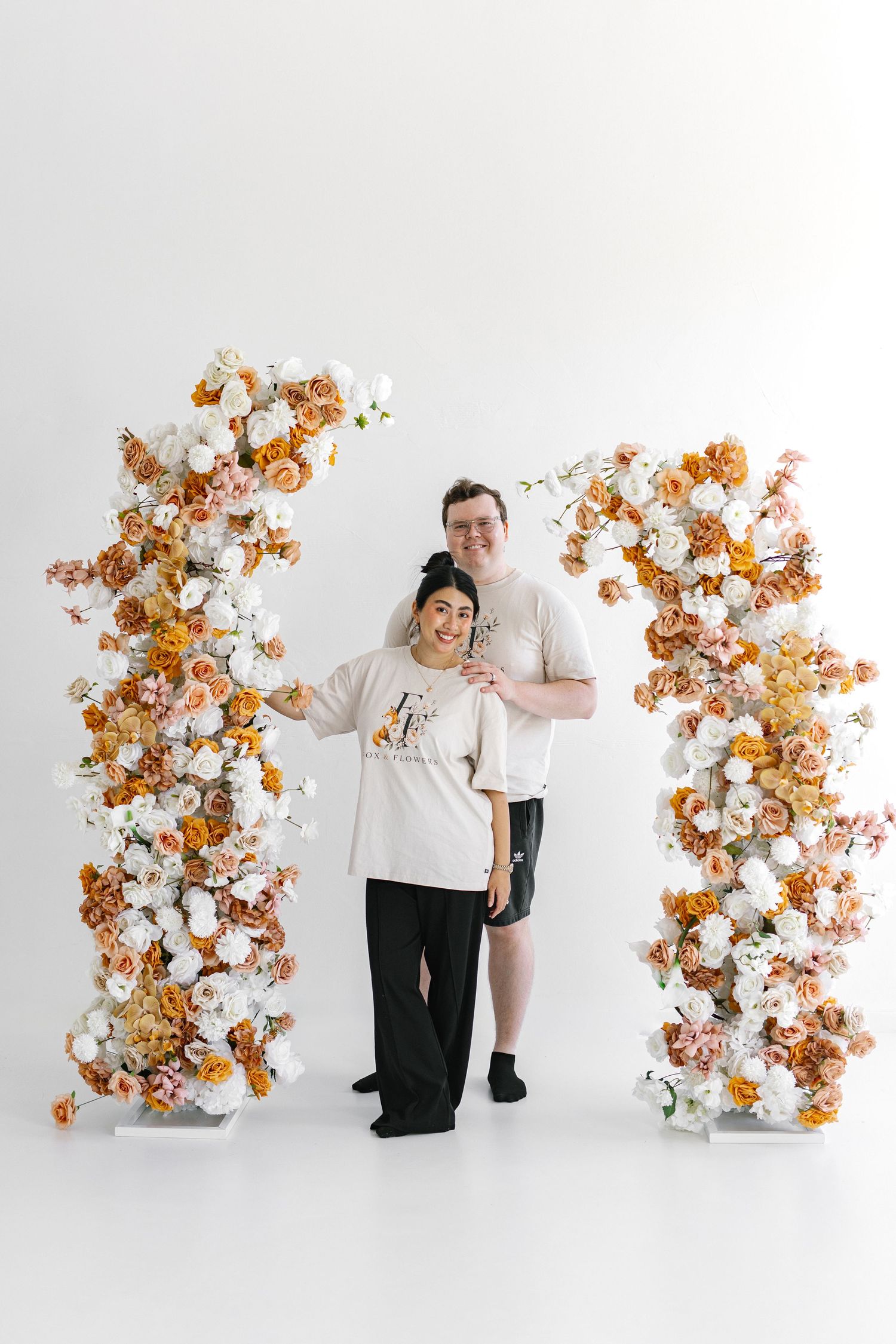 Couple smiling between two tall floral columns with white and orange roses on a white background.