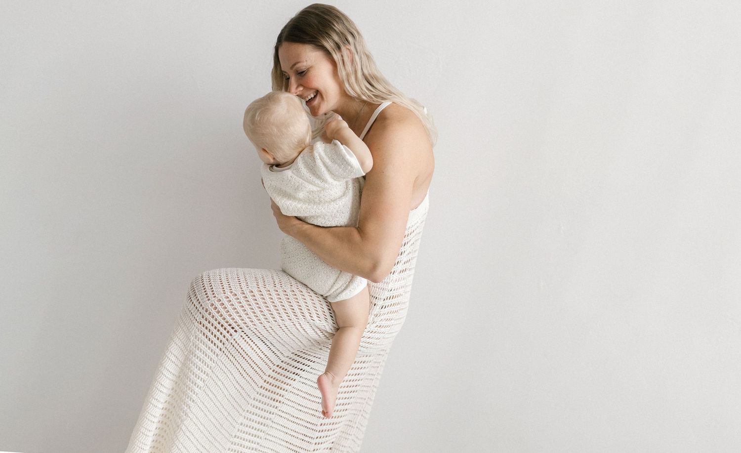 Smiling woman in a striped white maxi dress lovingly holds a baby in her arms against a clean, bright neutral studio background.