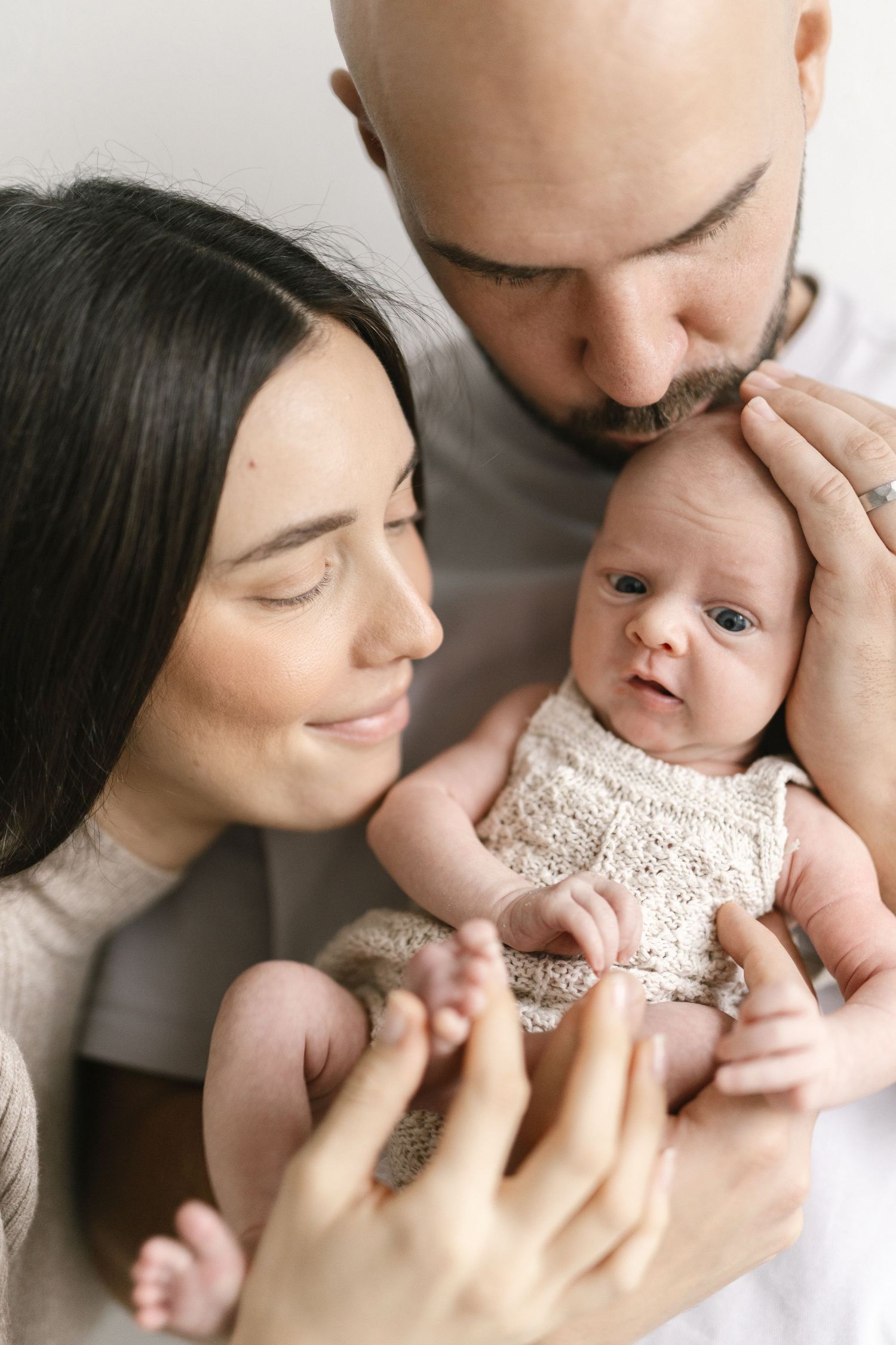 Smiling mother and bearded father gently cradle their newborn baby in a cream lace romper during a soft, intimate family portrait.