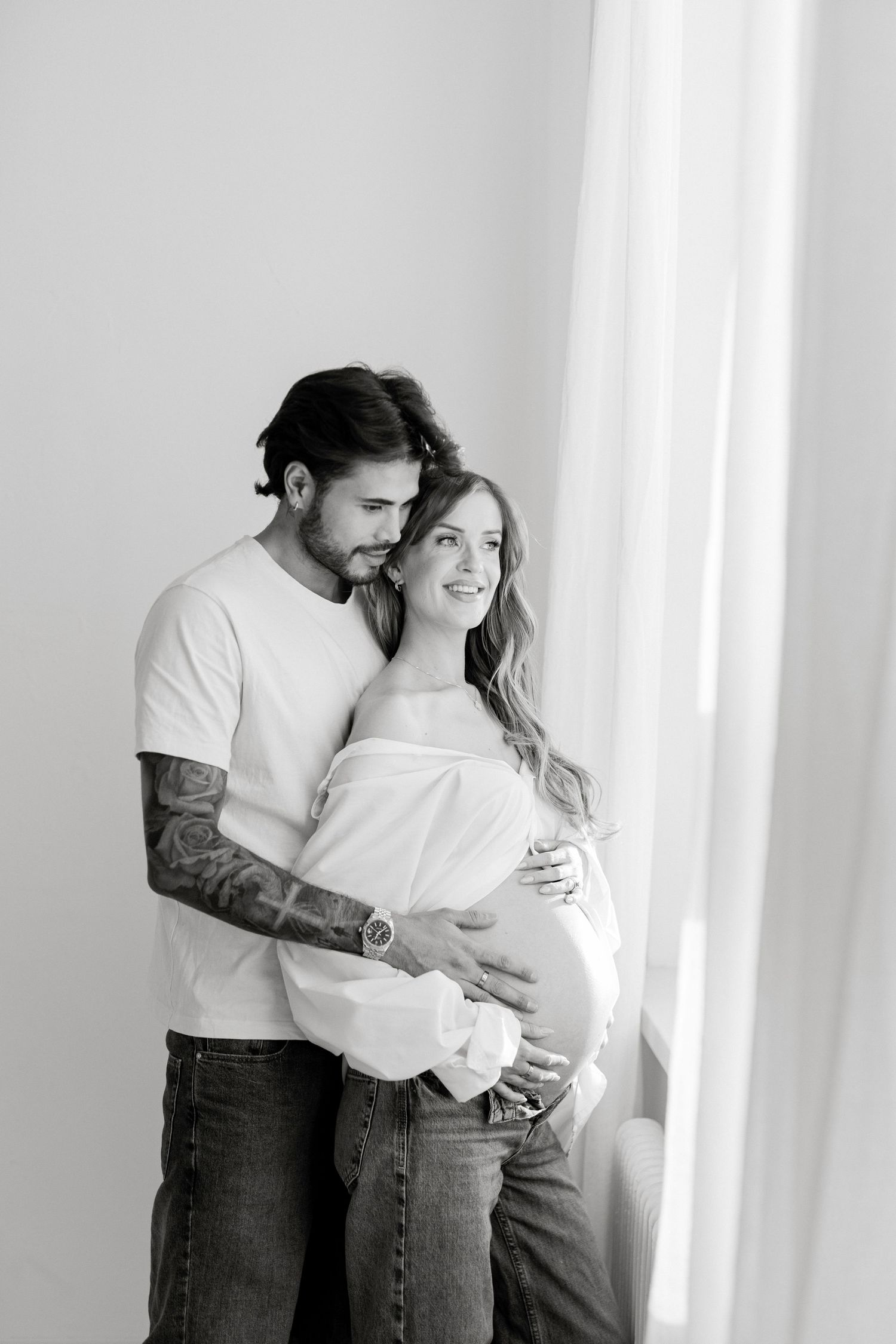Black and white maternity photo of a couple embracing, man with tattoos holding pregnant woman's belly near a window.
