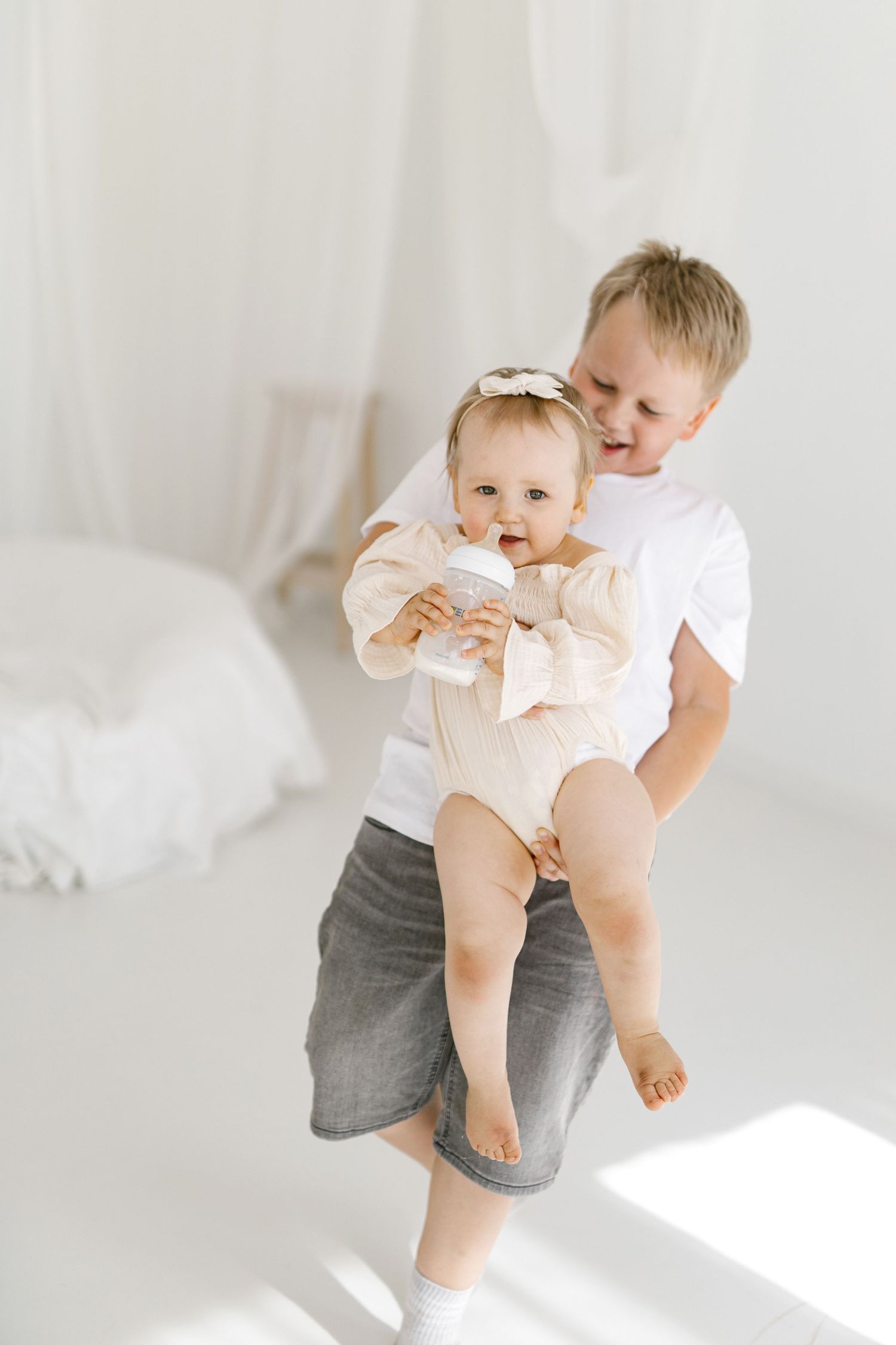 A smiling boy holds a baby girl in a white room, as she drinks from a bottle wearing a beige onesie and bow headband.