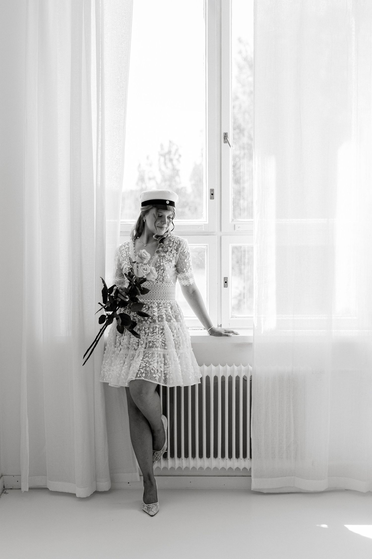 Bride in short white lace dress and cap holding bouquet by window in black and white photo.