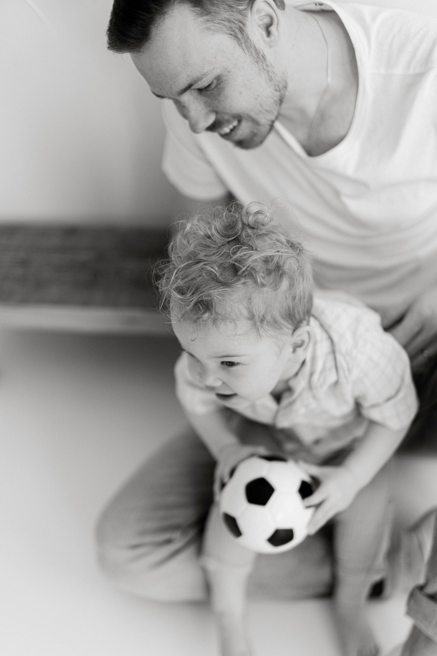 Black and white photo of a smiling father sitting with his curly-haired toddler holding a small soccer ball.