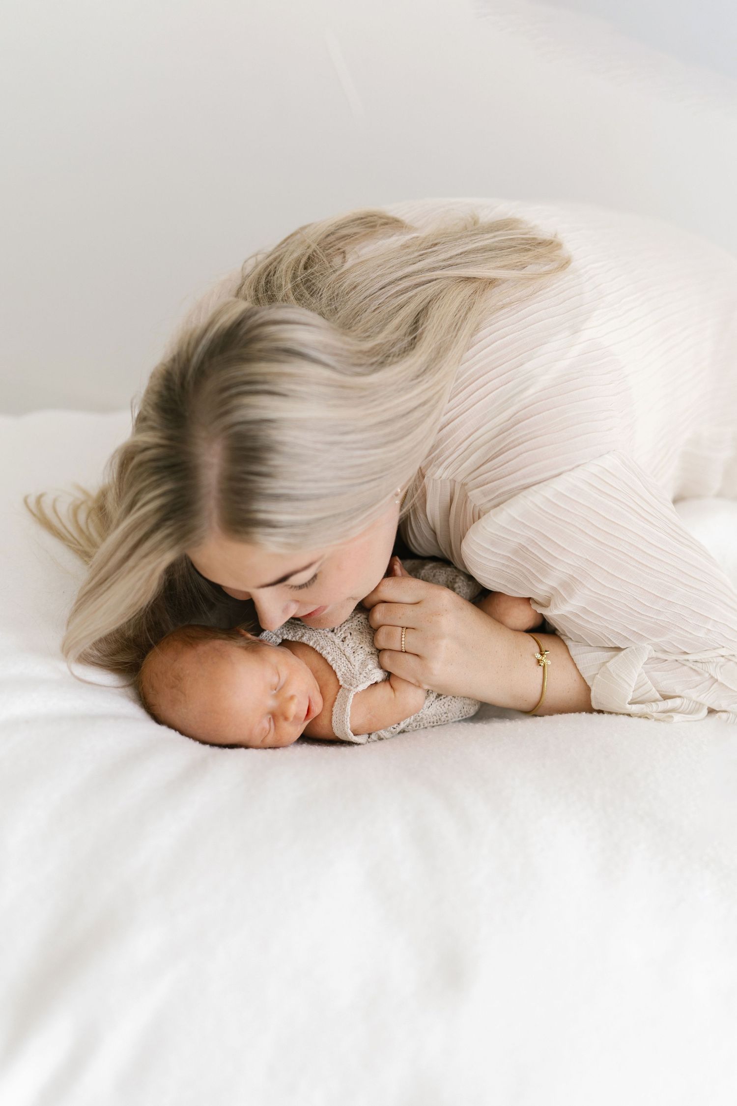 Mother tenderly kissing her sleeping newborn baby on a white bed, captured in a soft, bright newborn photography session.