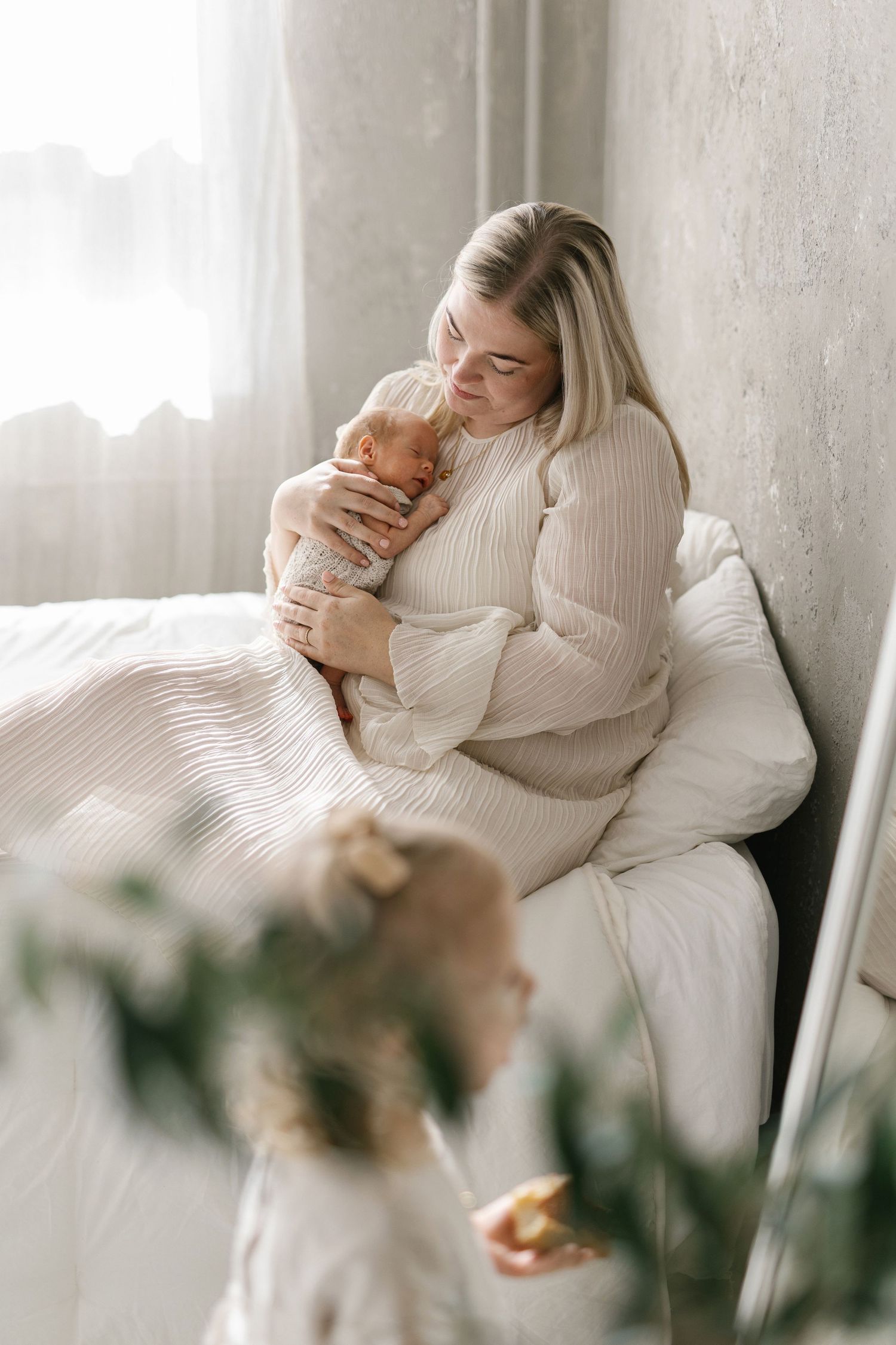 A blonde mother tenderly holds her newborn baby while sitting on a bed, as a young child stands nearby.
