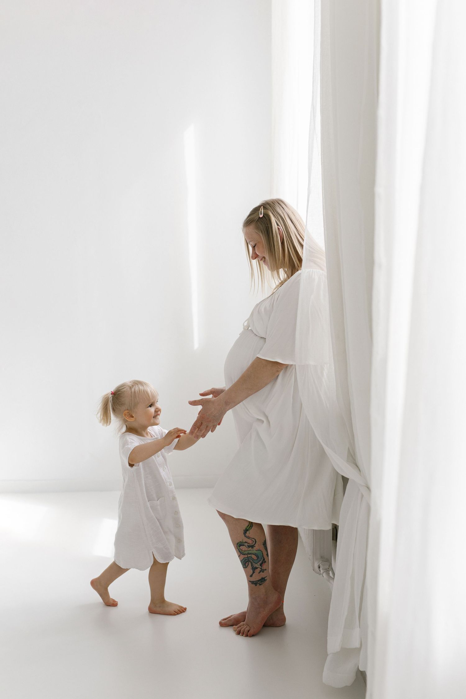 Pregnant mother in white robe holding hands with toddler daughter by white curtains.