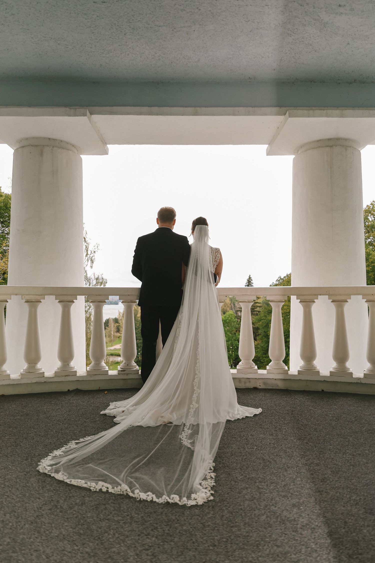 Bride and groom stand at elegant white balustrade with columns, bride wearing long lace cathedral veil trailing behind her.