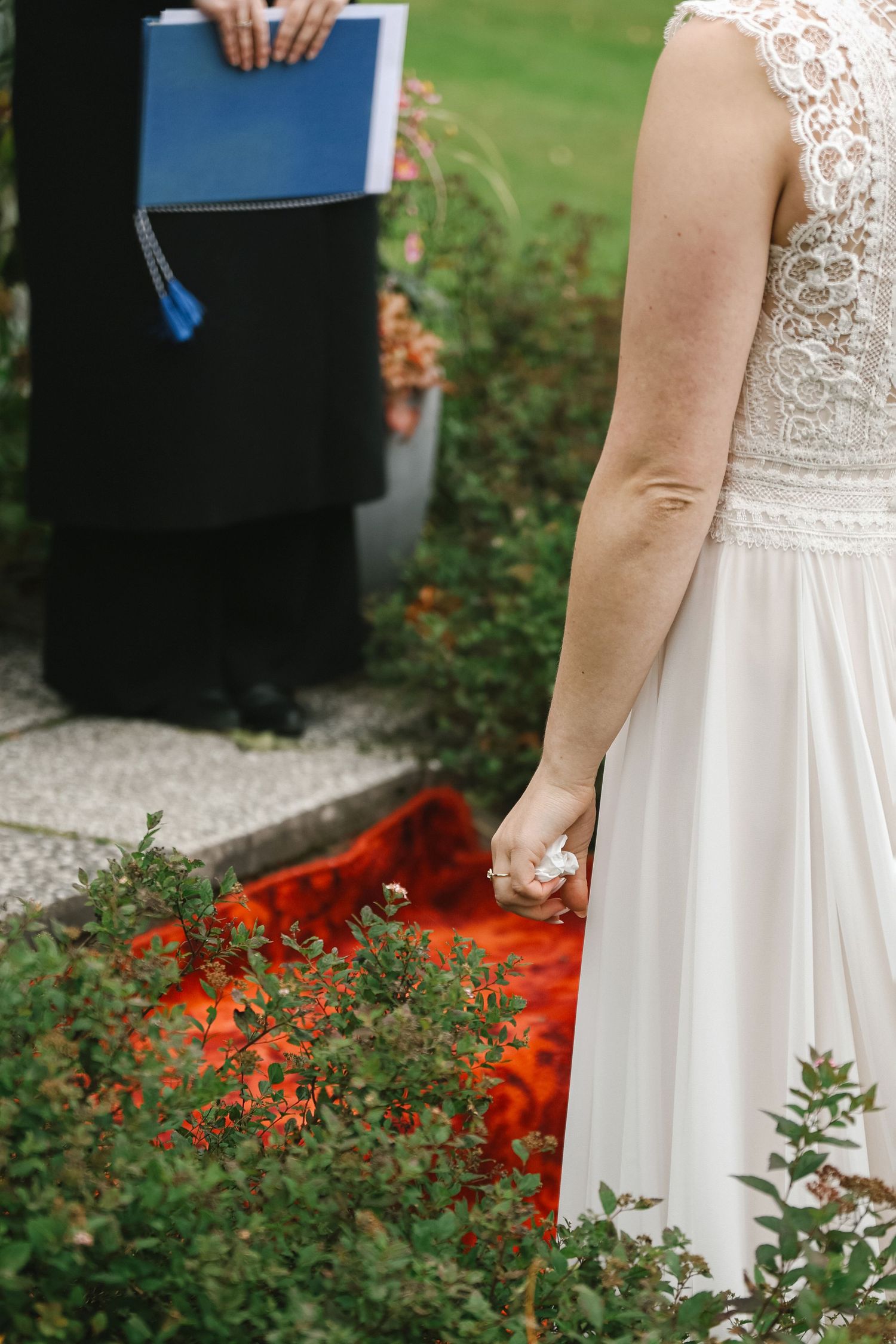 Bride in lace white gown stands near officiant holding blue book during outdoor wedding ceremony.