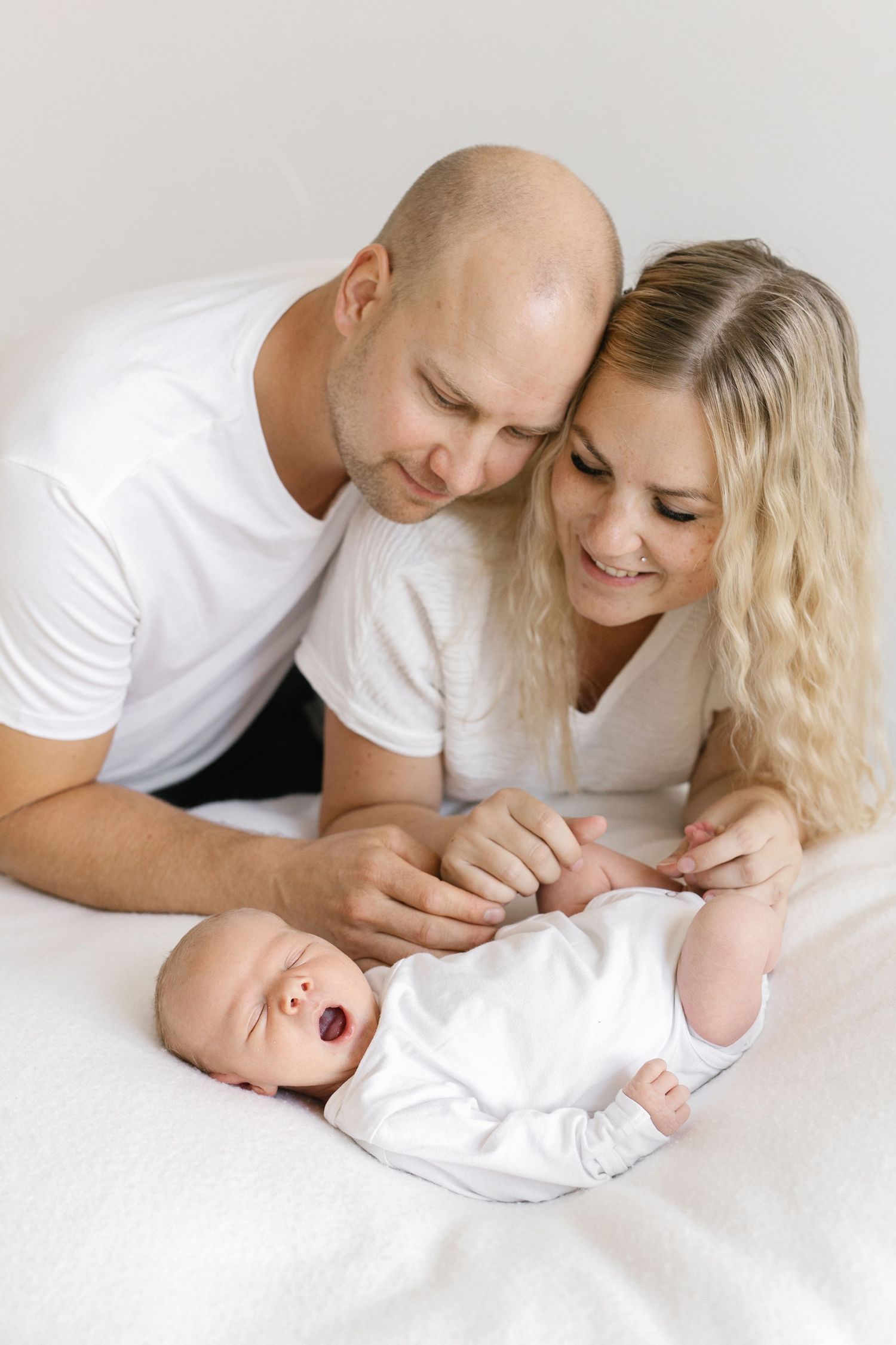A happy couple in white shirts tenderly gazes at their newborn baby lying on a white bed.