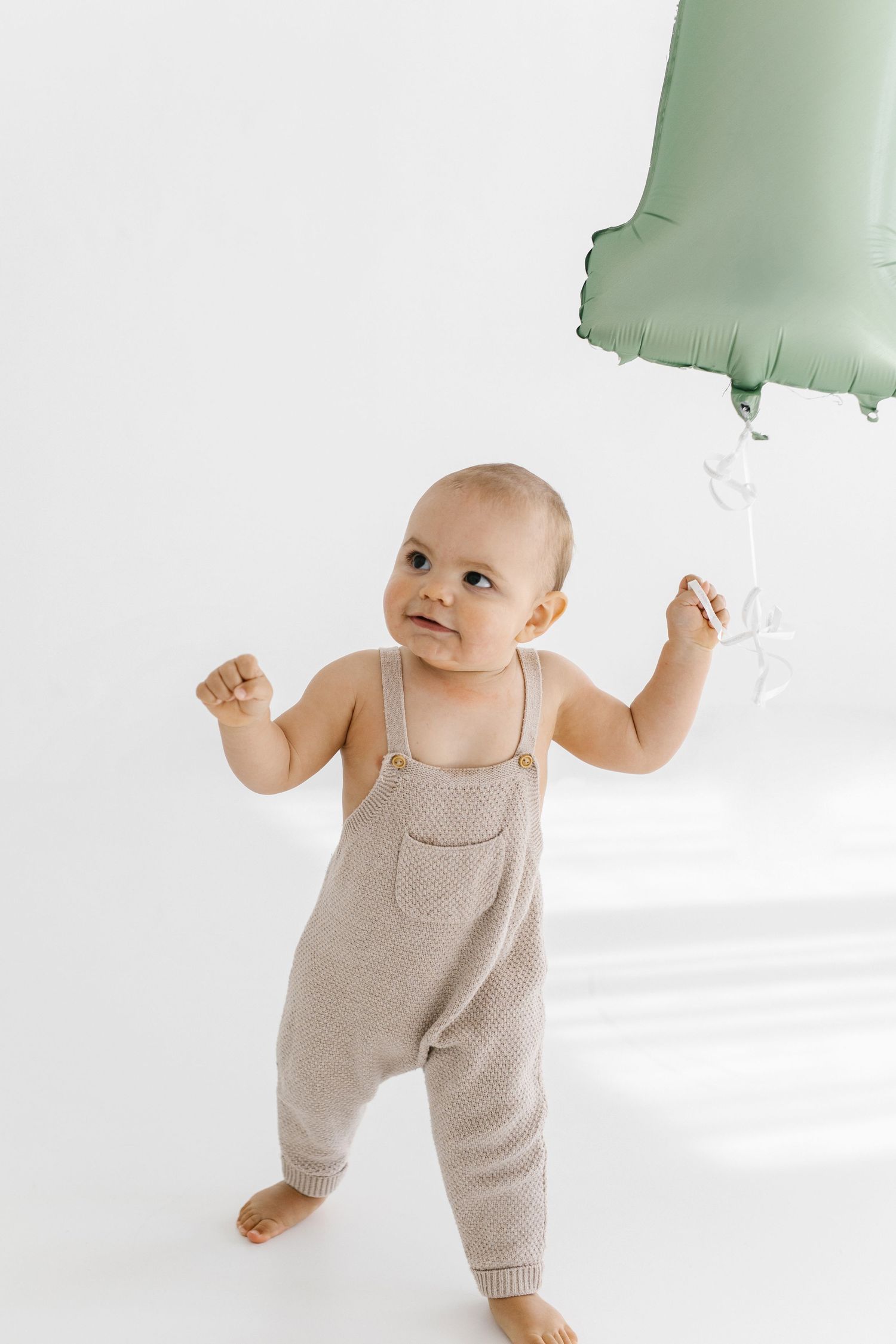 Baby in knit romper reaches for mint green balloon against white background.