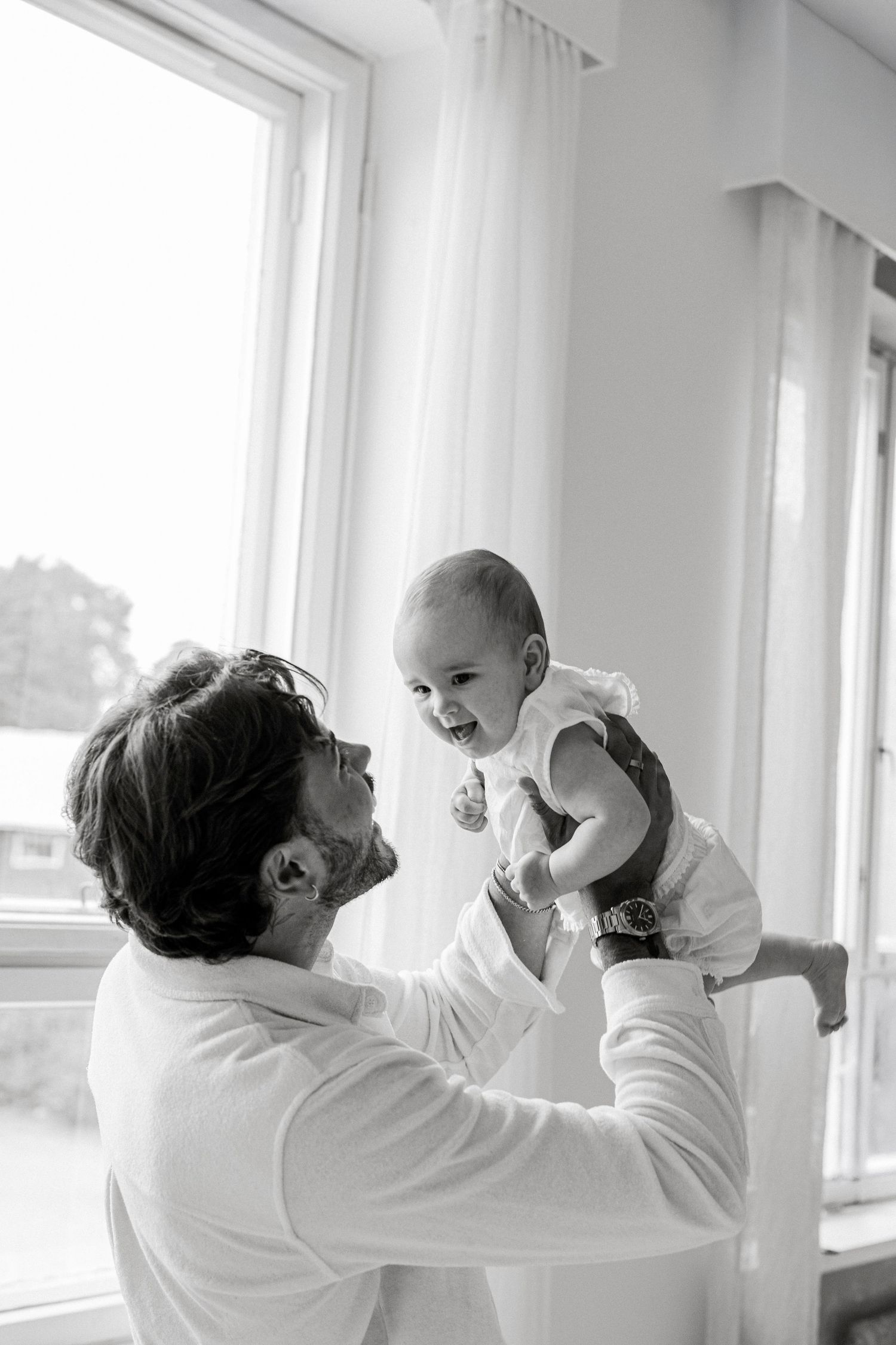 Black and white photo of a smiling man lifting a happy laughing baby near bright windows.