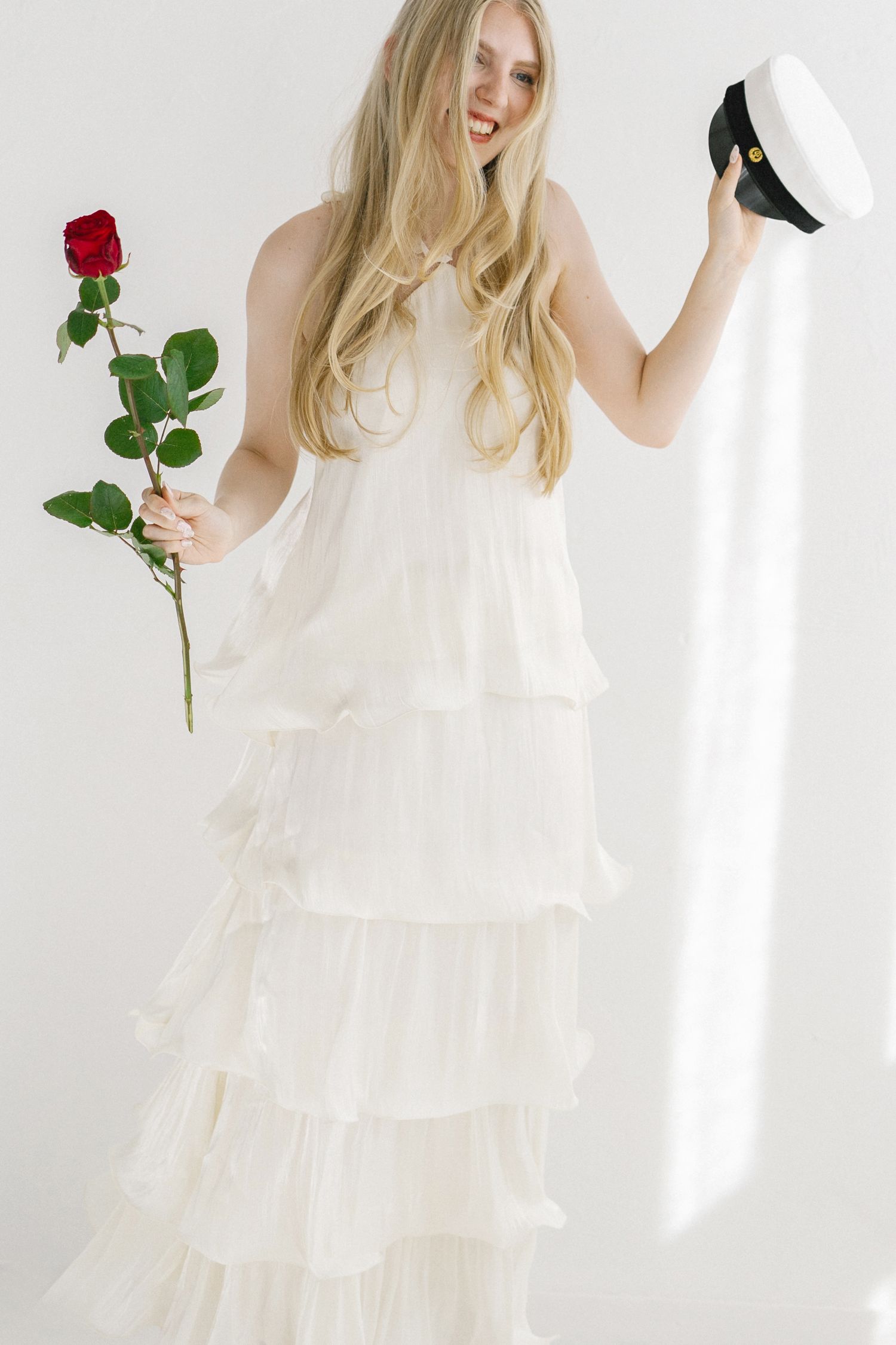 Smiling blonde woman in white tiered ruffle gown holding red rose and sailor hat against white background.