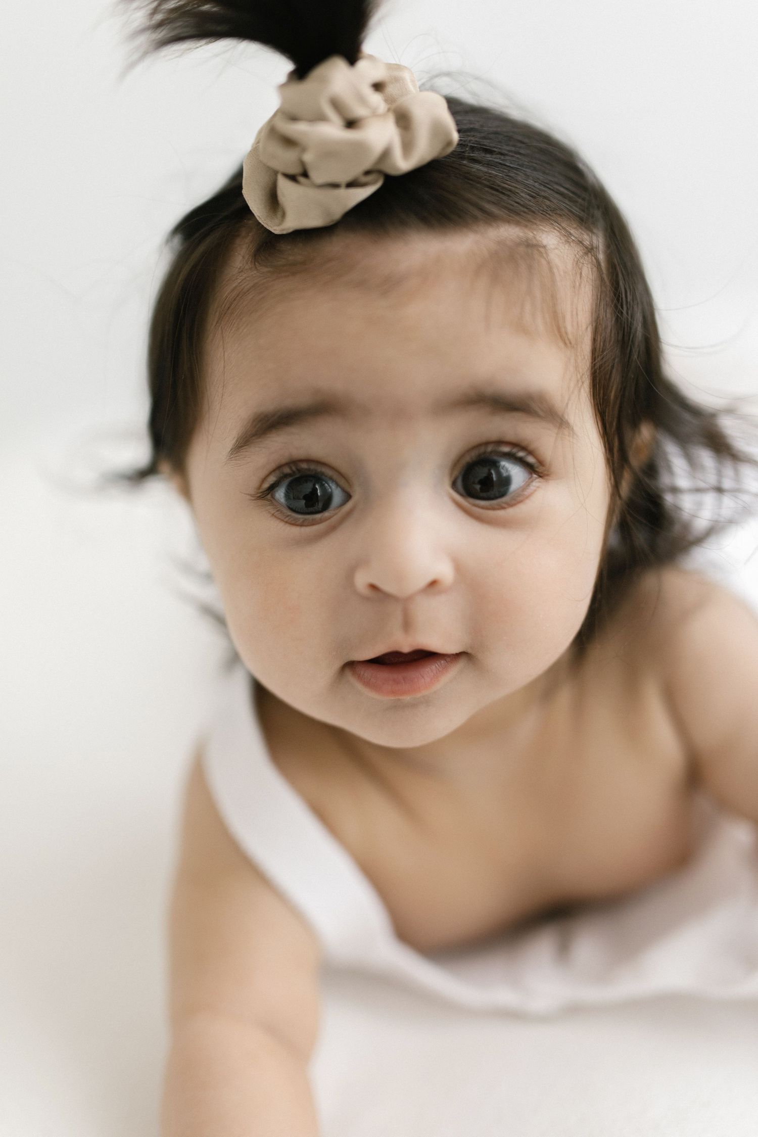 Adorable baby girl with dark hair and a beige bow scrunchie gazing up with big blue eyes.