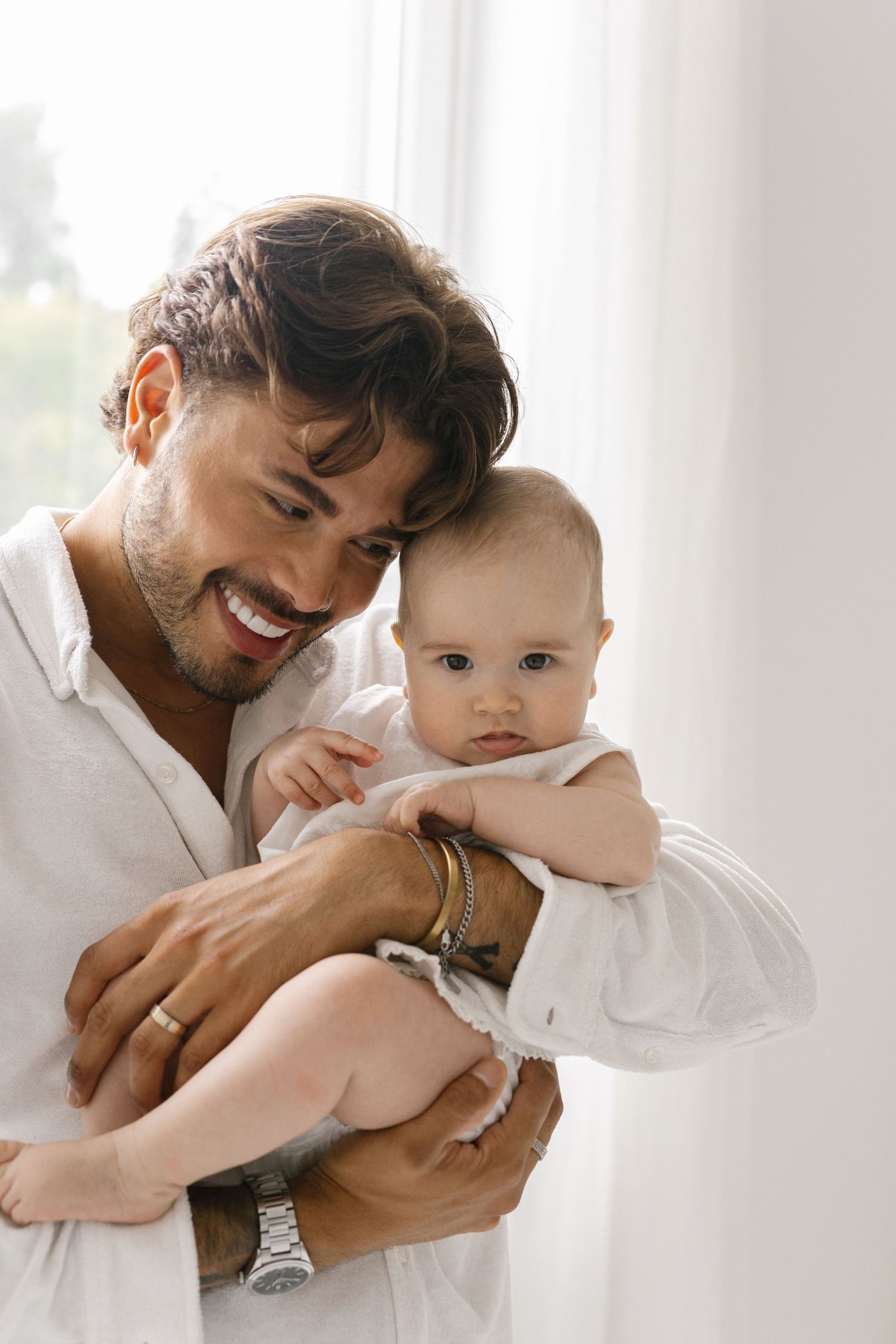 Smiling bearded man in a white linen shirt gently holds a baby in a white outfit near a sunlit window with sheer curtains.