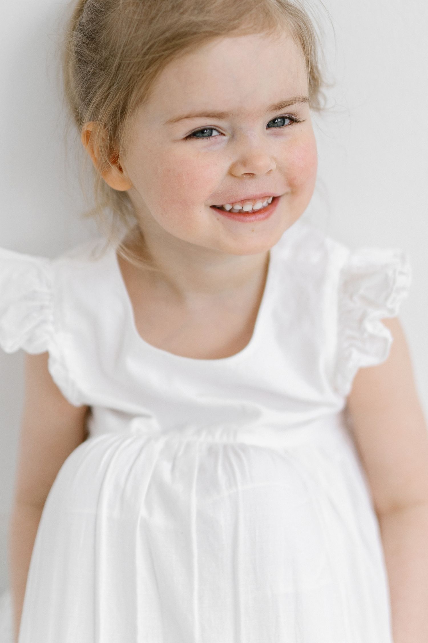 Smiling toddler girl with light brown hair wearing a white dress with ruffled flutter sleeves, leaning against a plain white wall.