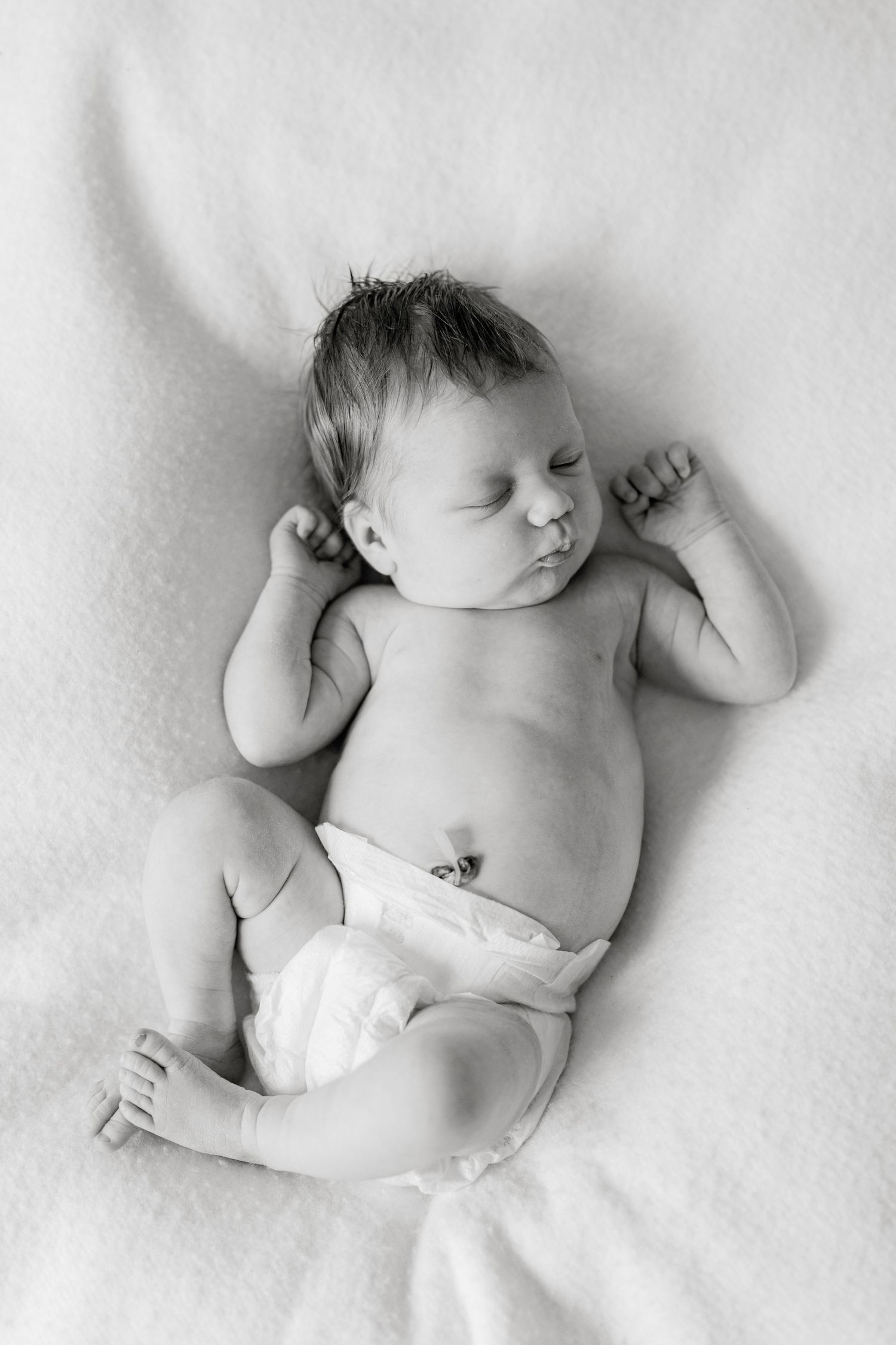 Black and white photo of a sleeping newborn baby in a diaper lying on a soft blanket with arms raised.