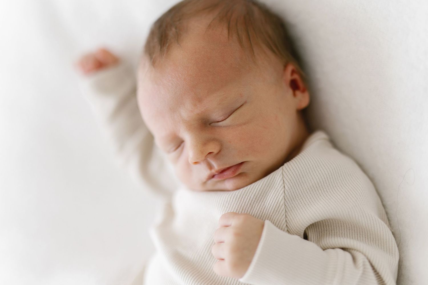 Peaceful newborn baby sleeping in white onesie on soft white blanket with tiny fist raised.