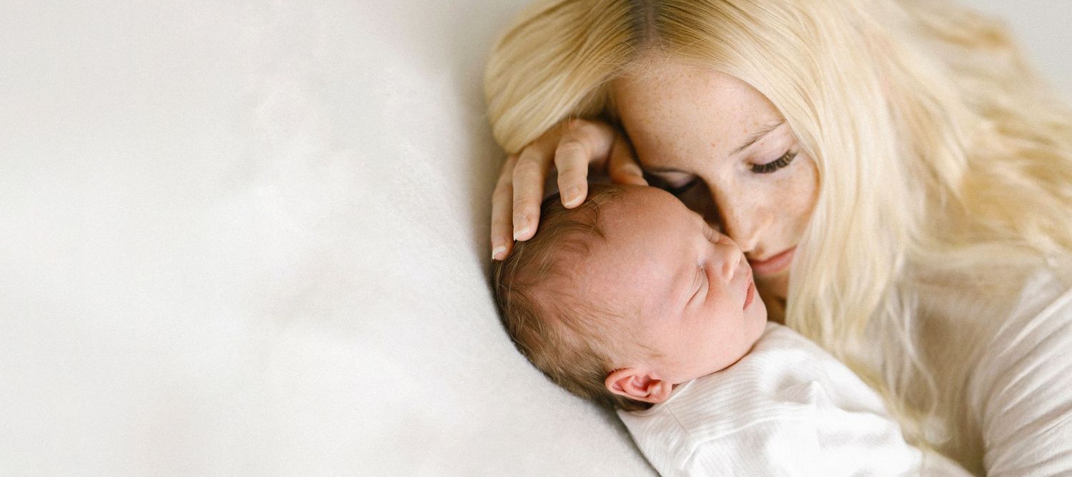 Tender moment of a blonde mother gently kissing her sleeping newborn baby wrapped in white.