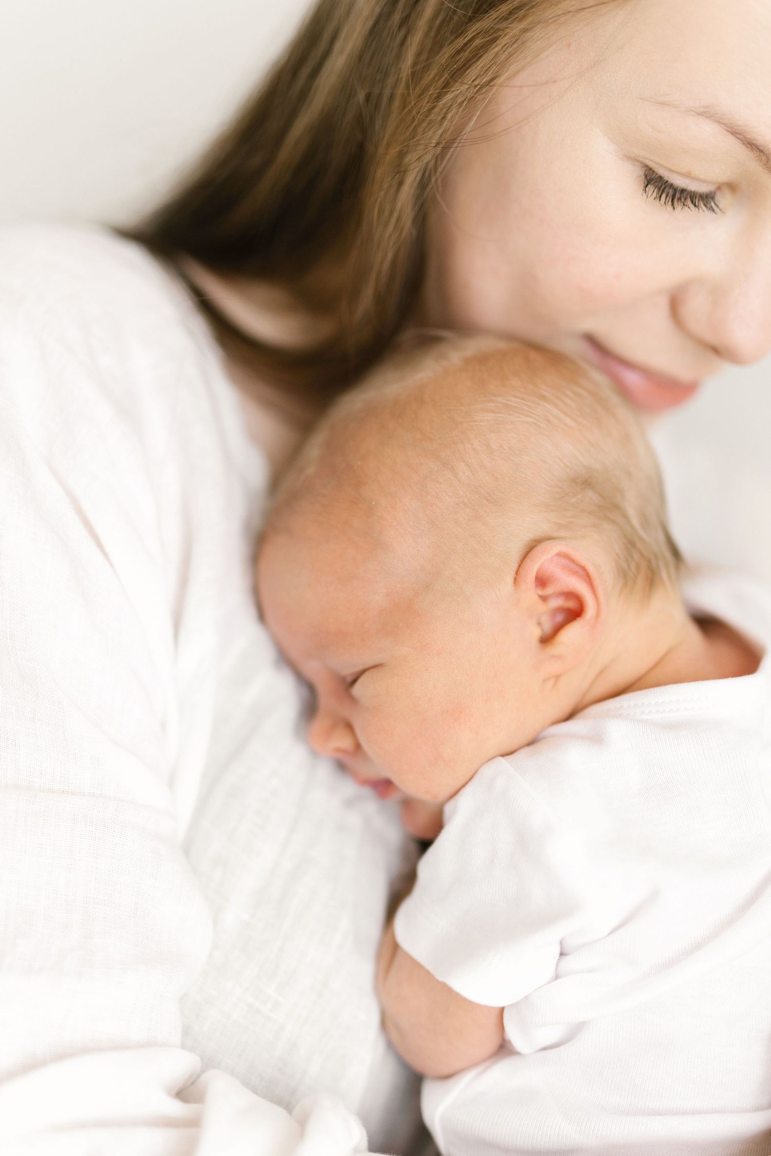 A mother in white gently cradles her sleeping newborn baby, both dressed in soft white clothing.