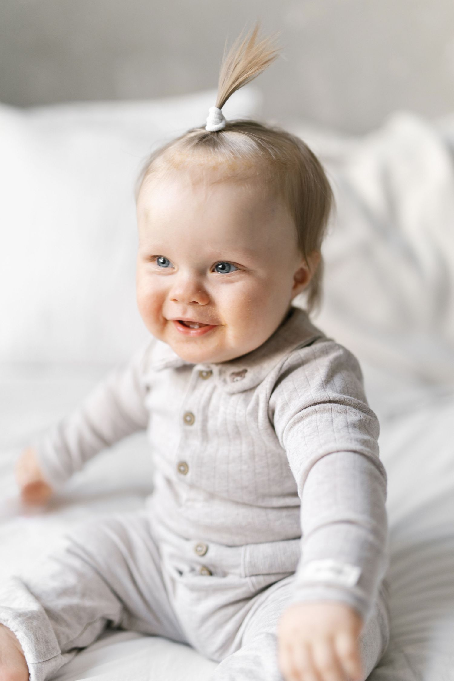 Baby in grey outfit smiling while lying on soft white bedding.