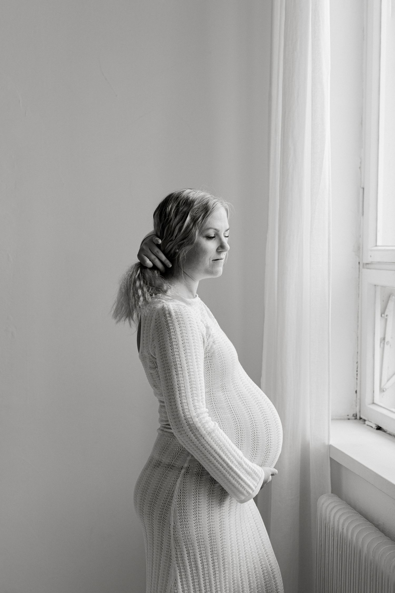 Black and white maternity portrait of a pregnant woman in a knit dress cradling her belly beside a softly lit window.