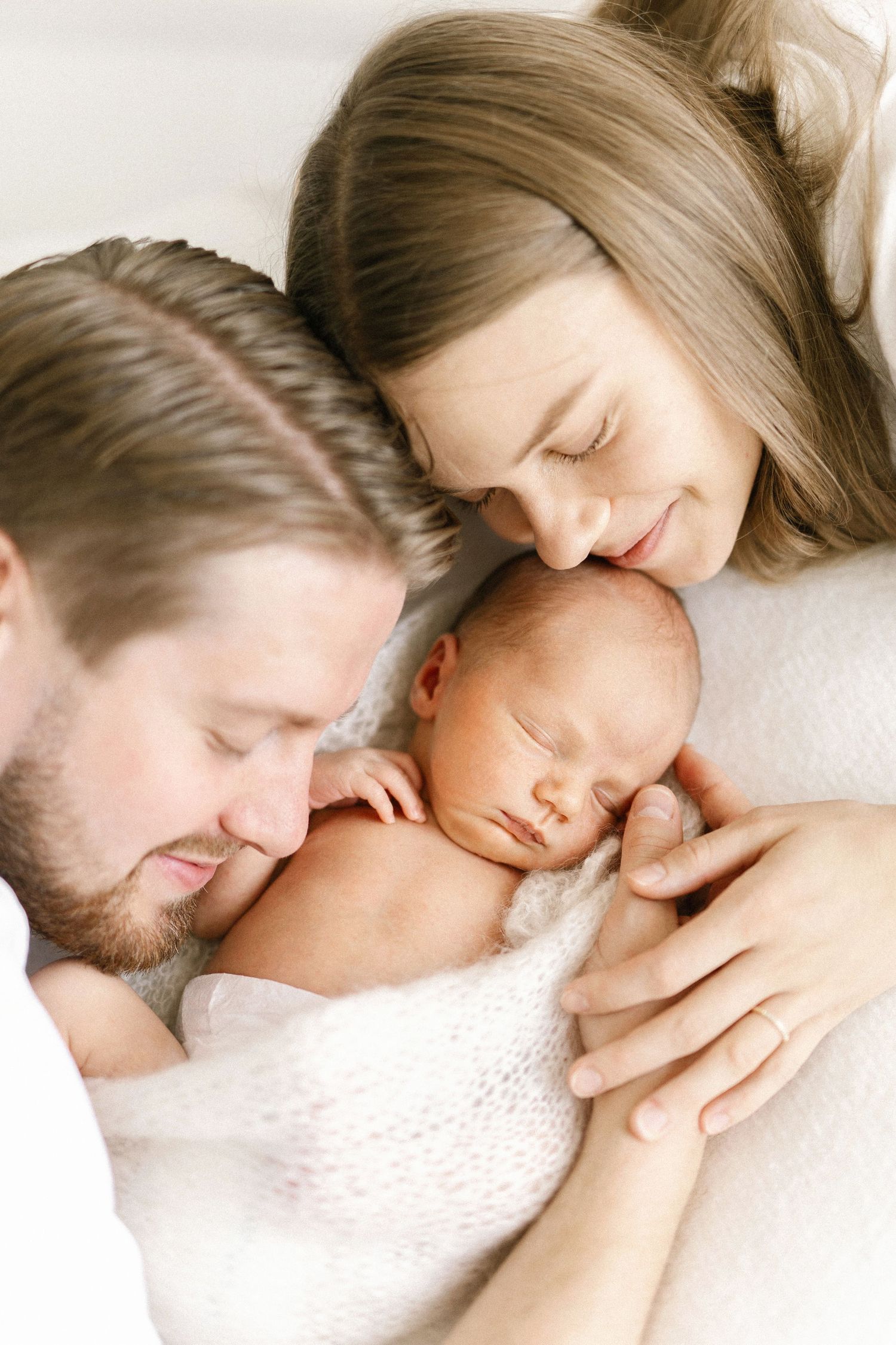 Parents gently lean over their sleeping newborn baby wrapped in a soft white blanket in a tender family portrait.