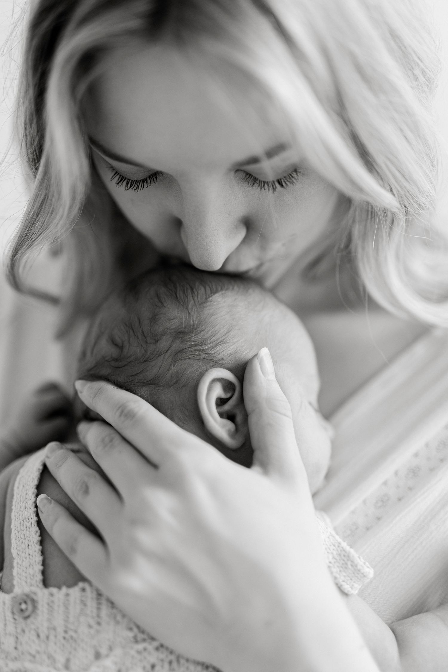Black and white photo of a mother tenderly kissing her newborn baby's head while cradling them gently.