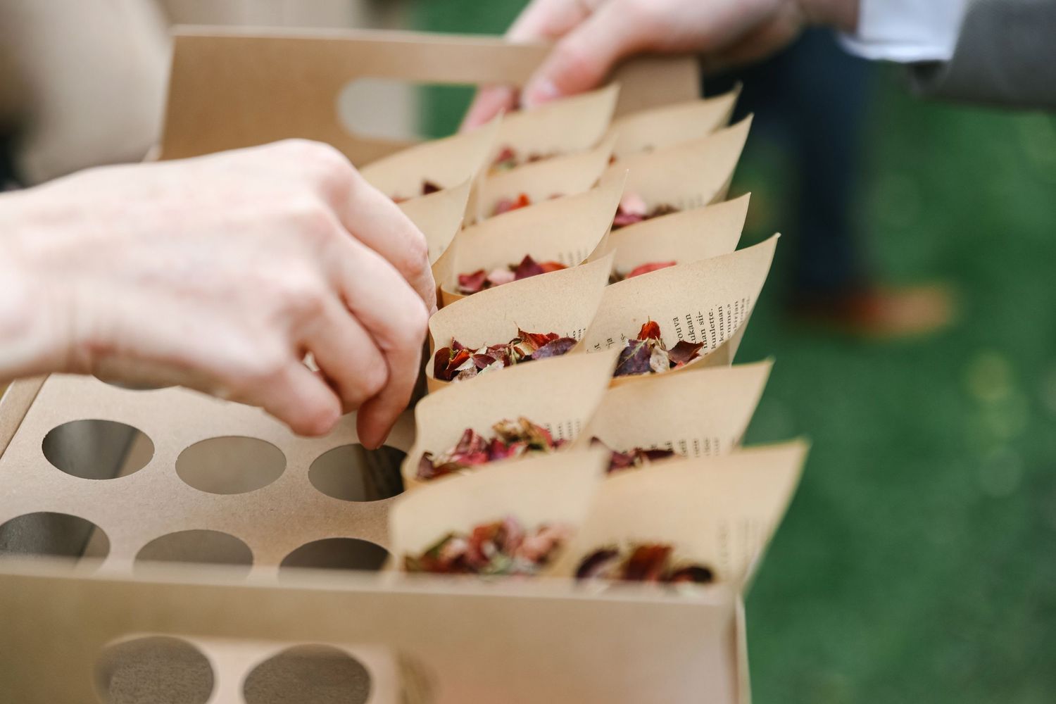 Hands arranging colorful floral confetti in cardboard wedding favor boxes displayed in a rustic holder outdoors.