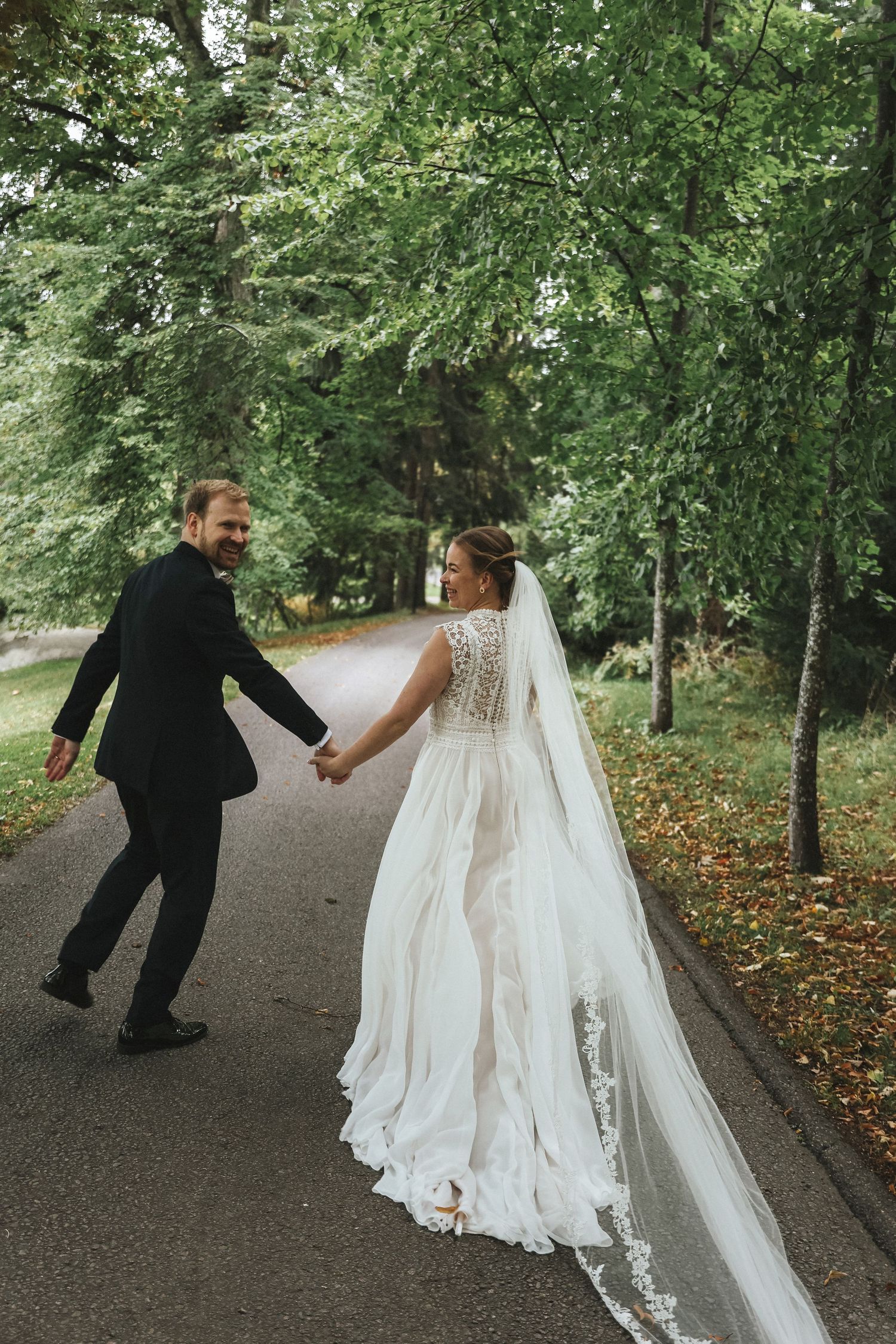 Bride and groom walk hand in hand along tree-lined path in lush green park on wedding day.