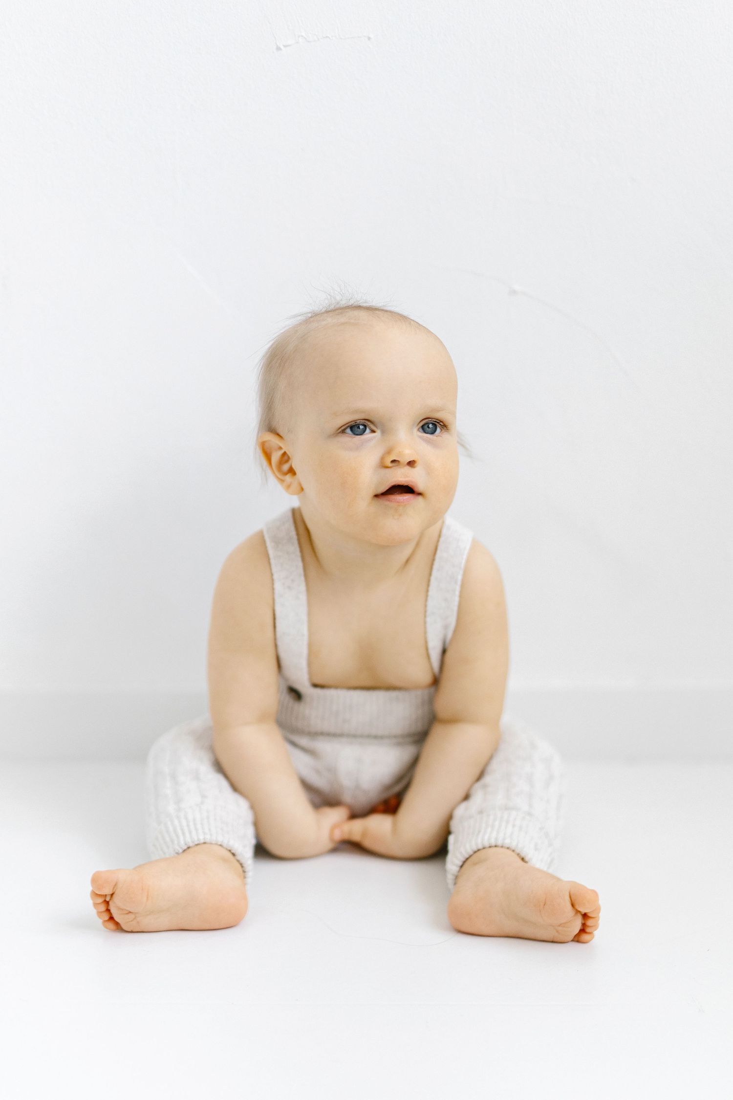 Adorable baby in white knit overalls sitting on a white floor, looking up with curious blue eyes.