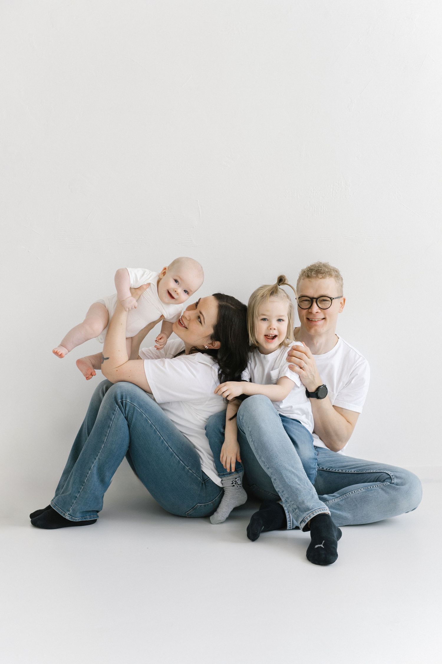 A family wearing jeans and white shirts poses playfully while lying down on a white studio backdrop.