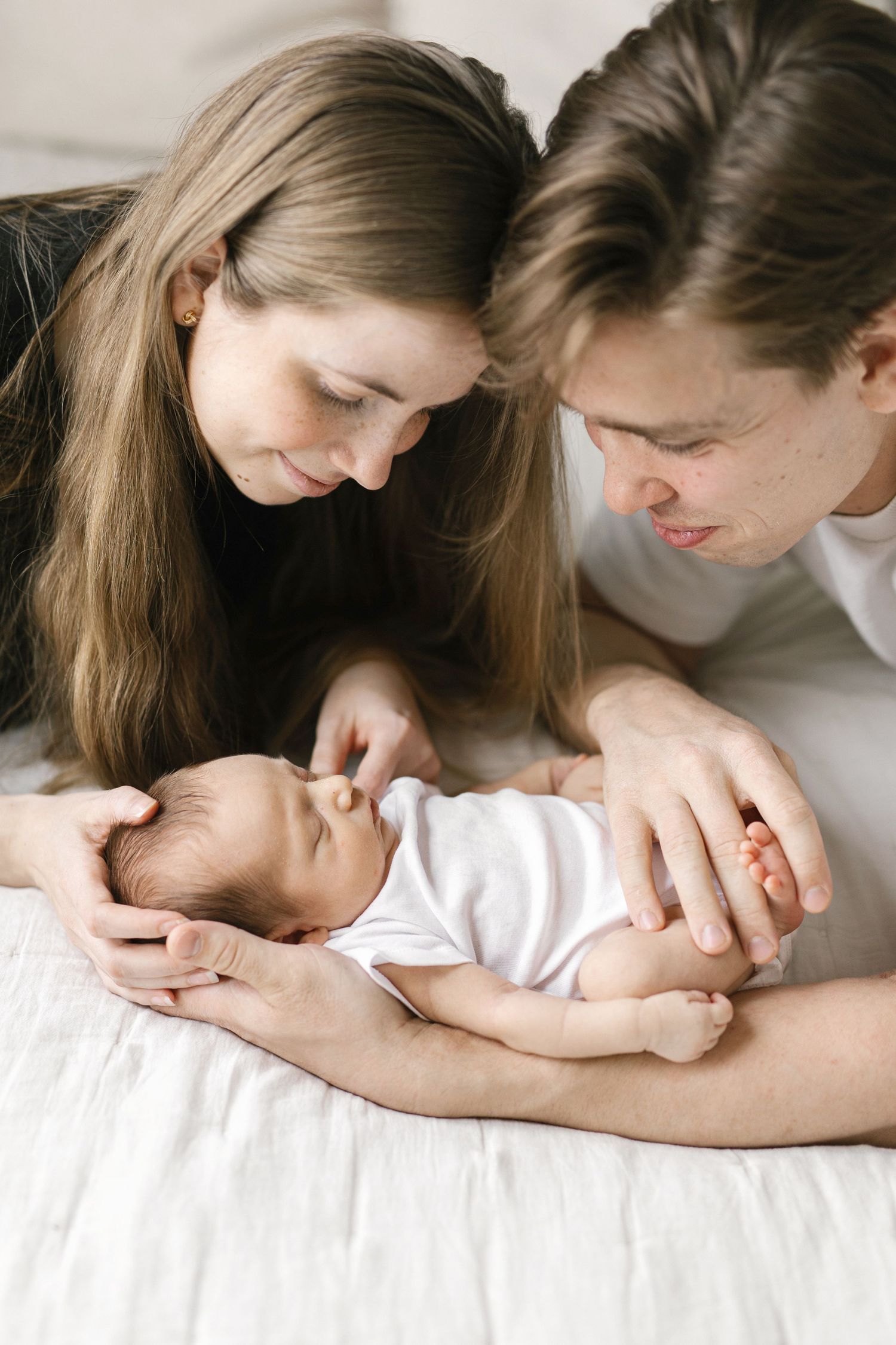Young parents lovingly gazing at their newborn baby lying on a white bed in a tender family moment.