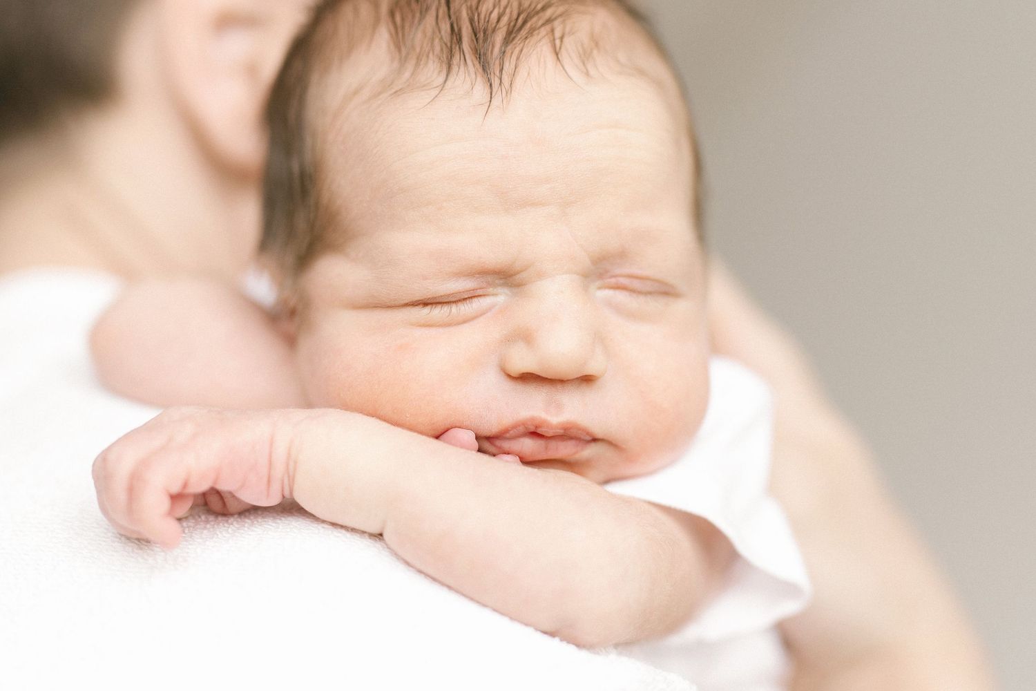 Sleeping newborn baby with scrunched face resting on an adult's shoulder, wrapped in soft white, in a gentle newborn portrait.