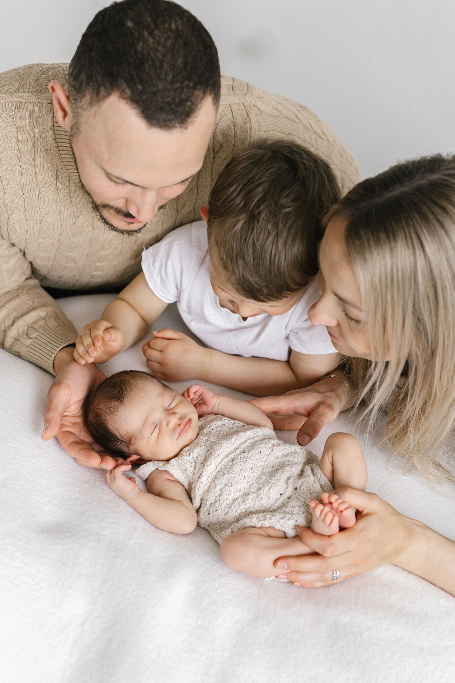 A loving family of four admires their sleeping newborn baby, with parents and toddler gathered closely on a white bed.