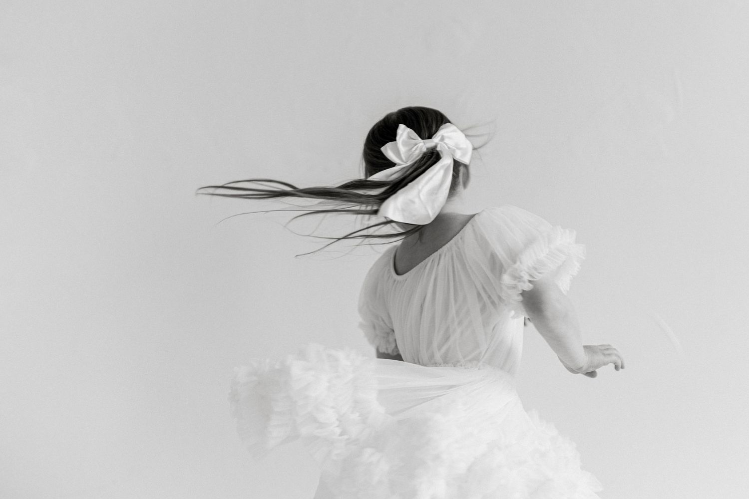 Black and white photo of a girl in a white ruffled dress, hair flowing with a bow, captured mid-twirl.