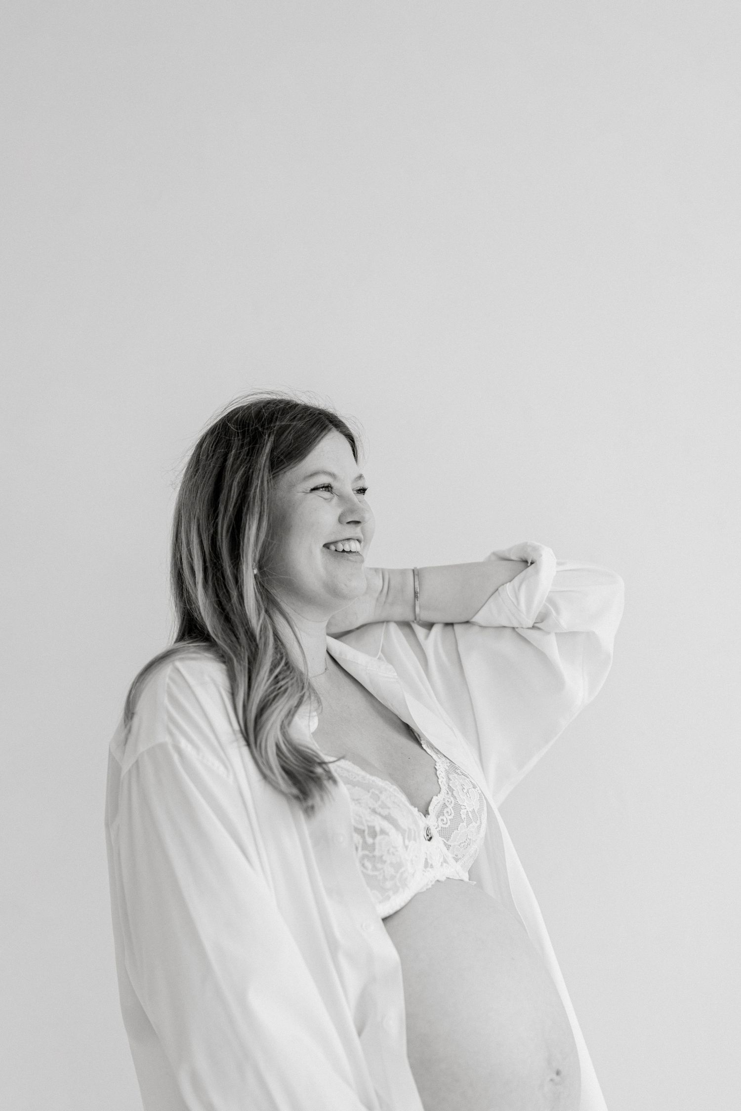 Black and white photo of a smiling pregnant woman in a white shirt and lace bra, posing against a light background.