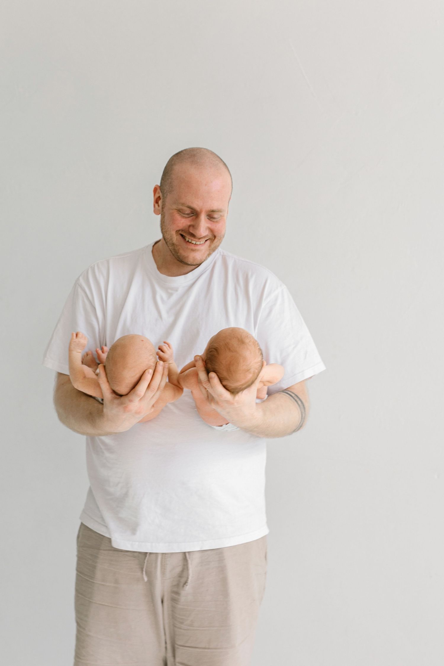 Smiling bald father holding two newborn babies, one in each arm, wearing a white t-shirt against a light background.