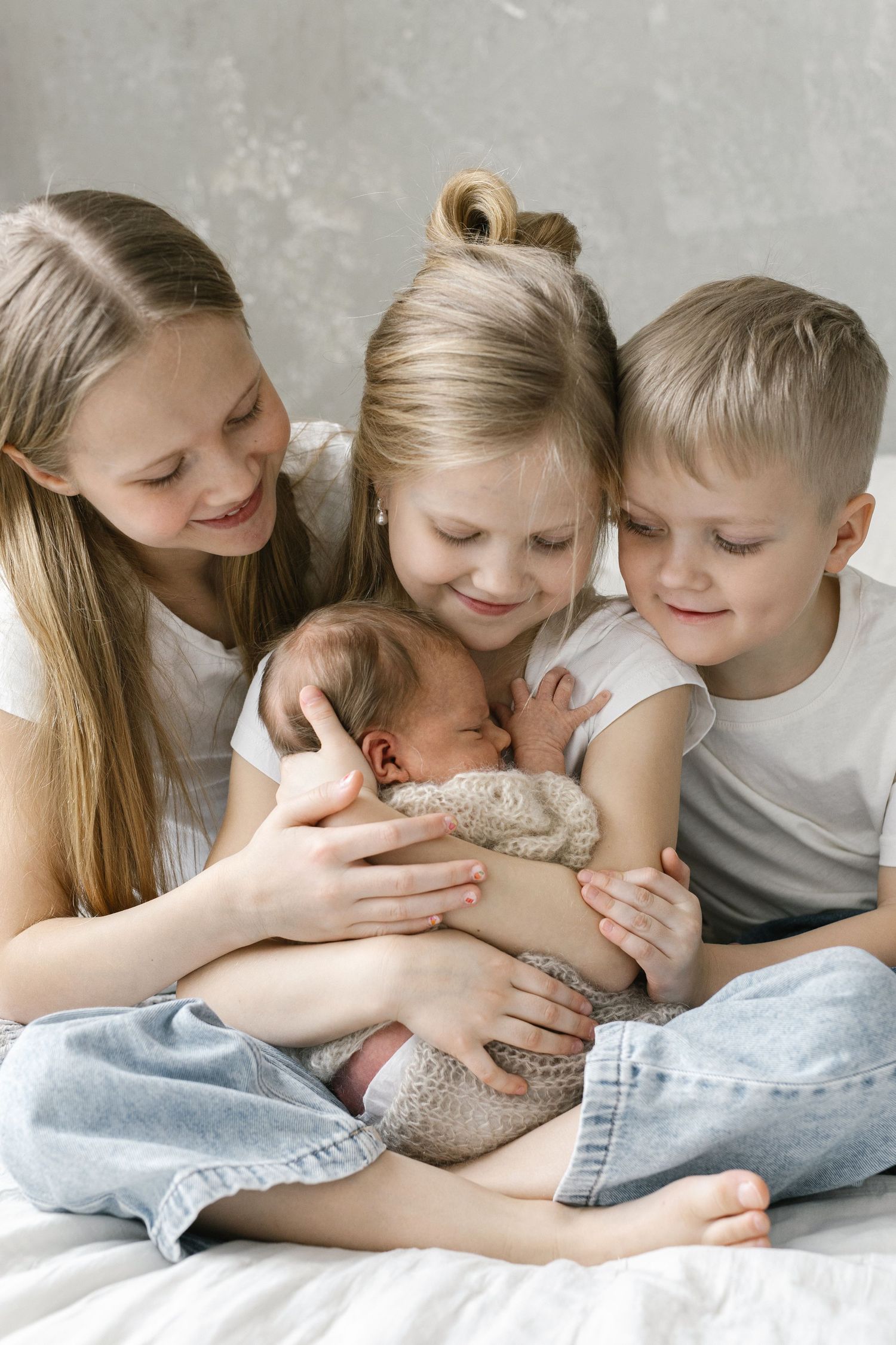 Three older siblings lovingly cradle their newborn baby together on a white bed.
