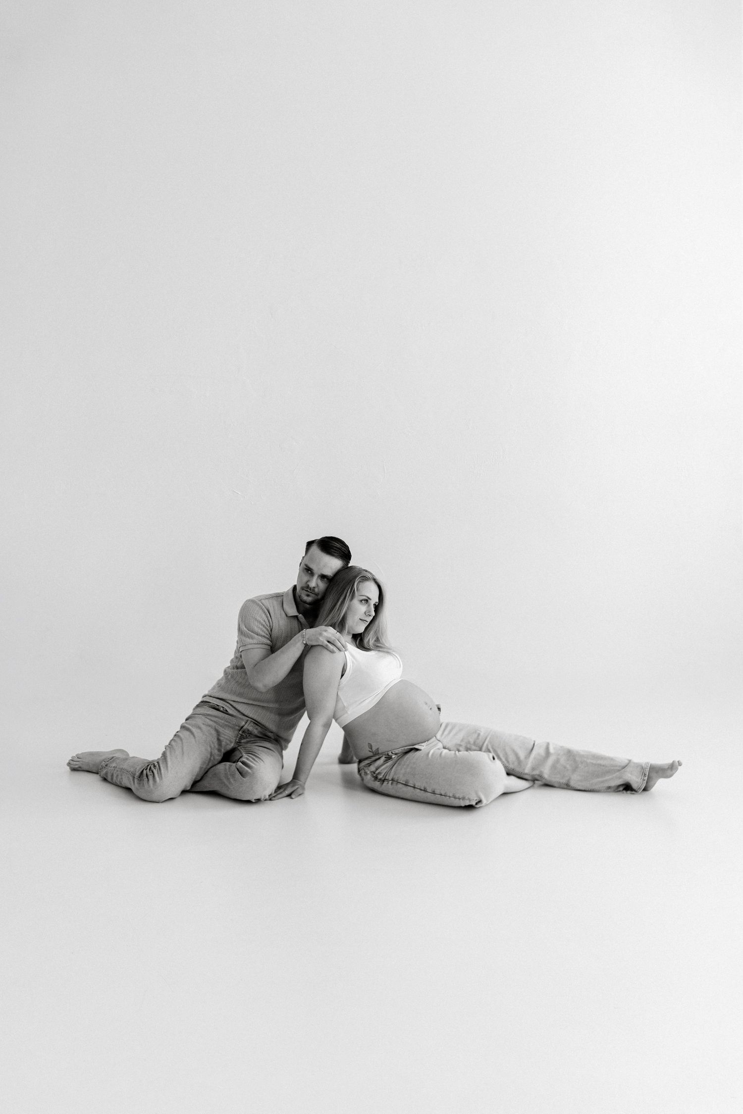 Black and white photo of a pregnant couple sitting on the floor, man embracing woman from behind in a studio.