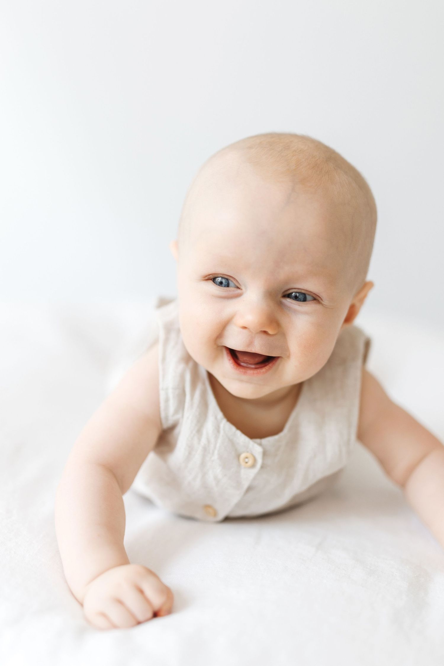 Smiling baby in a beige romper doing tummy time on a white surface, looking up with bright blue eyes.