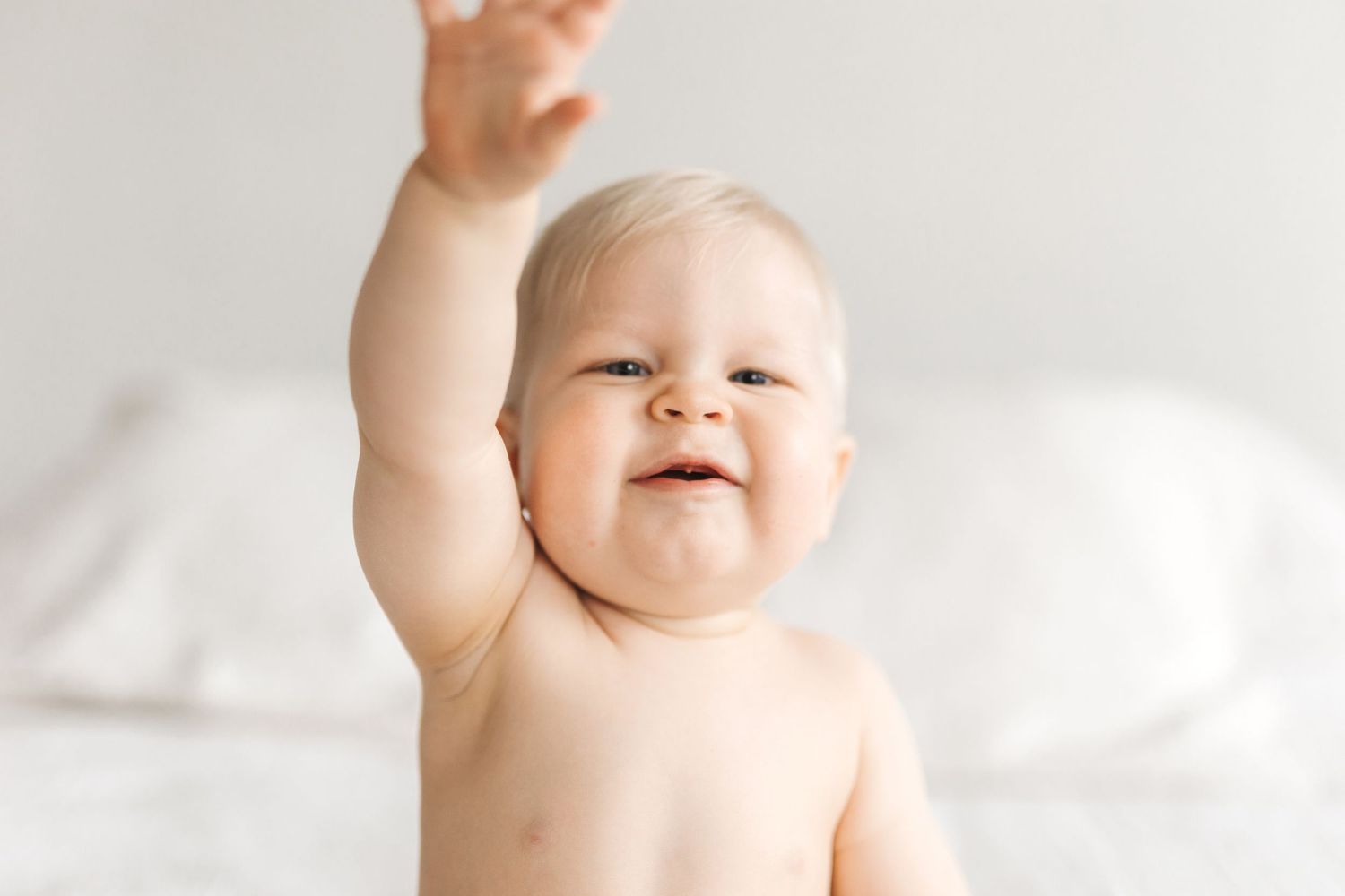 Happy baby raising one arm up high, smiling joyfully while sitting against a soft white background.