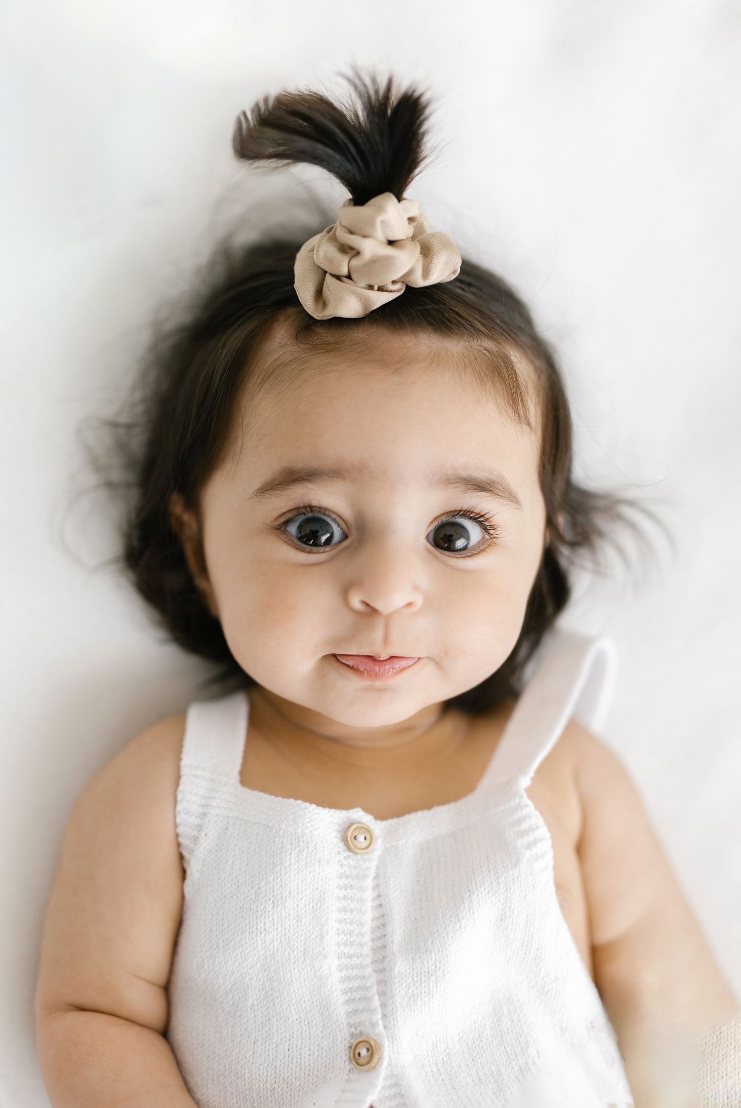 Cute baby girl lying down wearing white romper with beige floral scrunchie and expressive blue eyes.