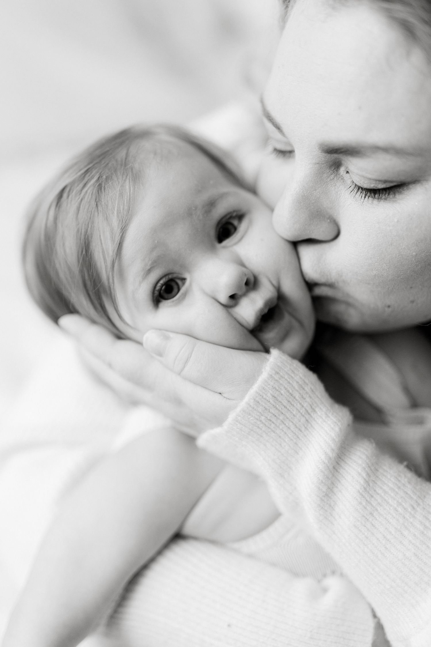 Black and white photo of a mother tenderly kissing her newborn baby's cheek while cradling them gently.